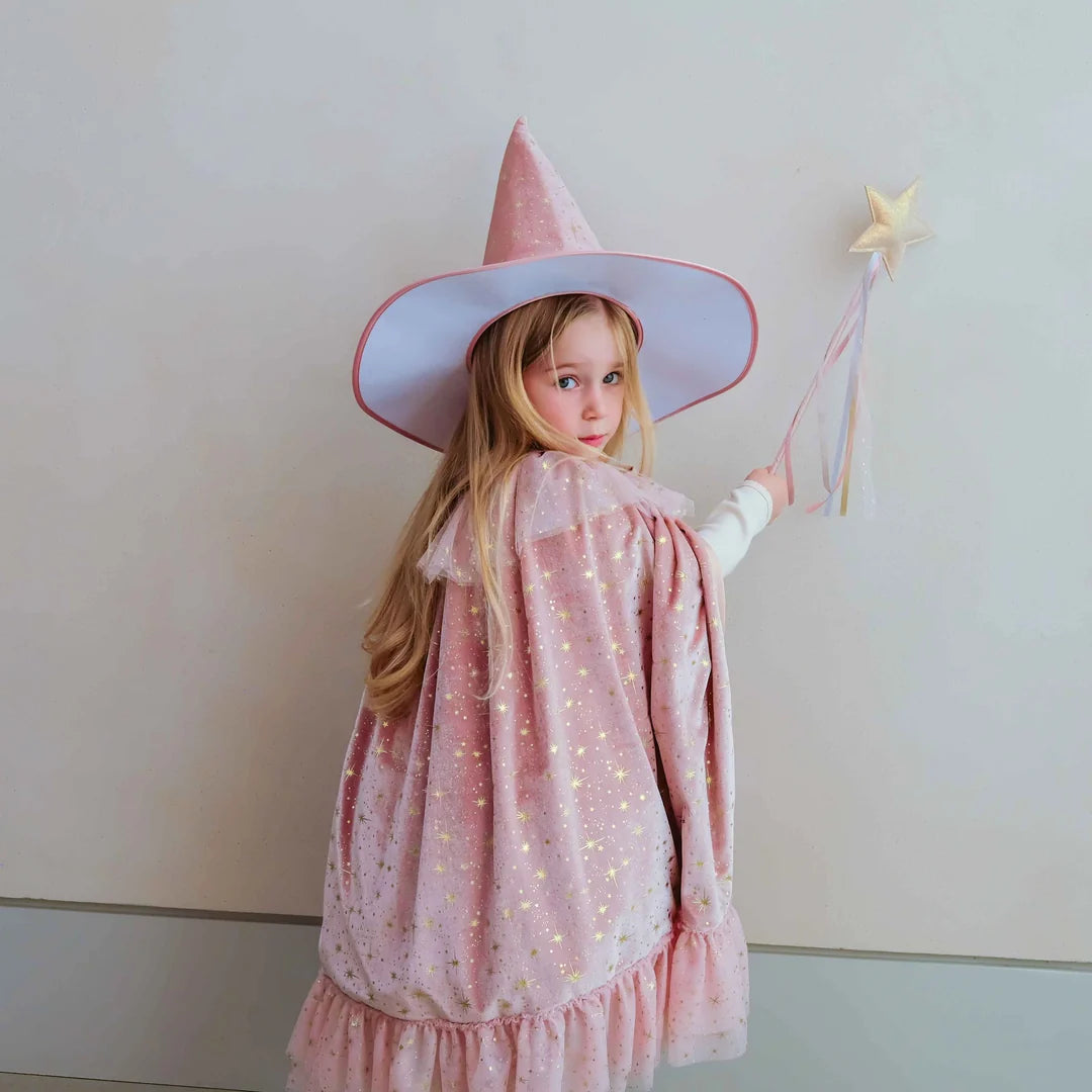 A young girl wearing a pink witch costume with a pointed hat, star wand, and sparkly cape, standing against a plain light beige wall.