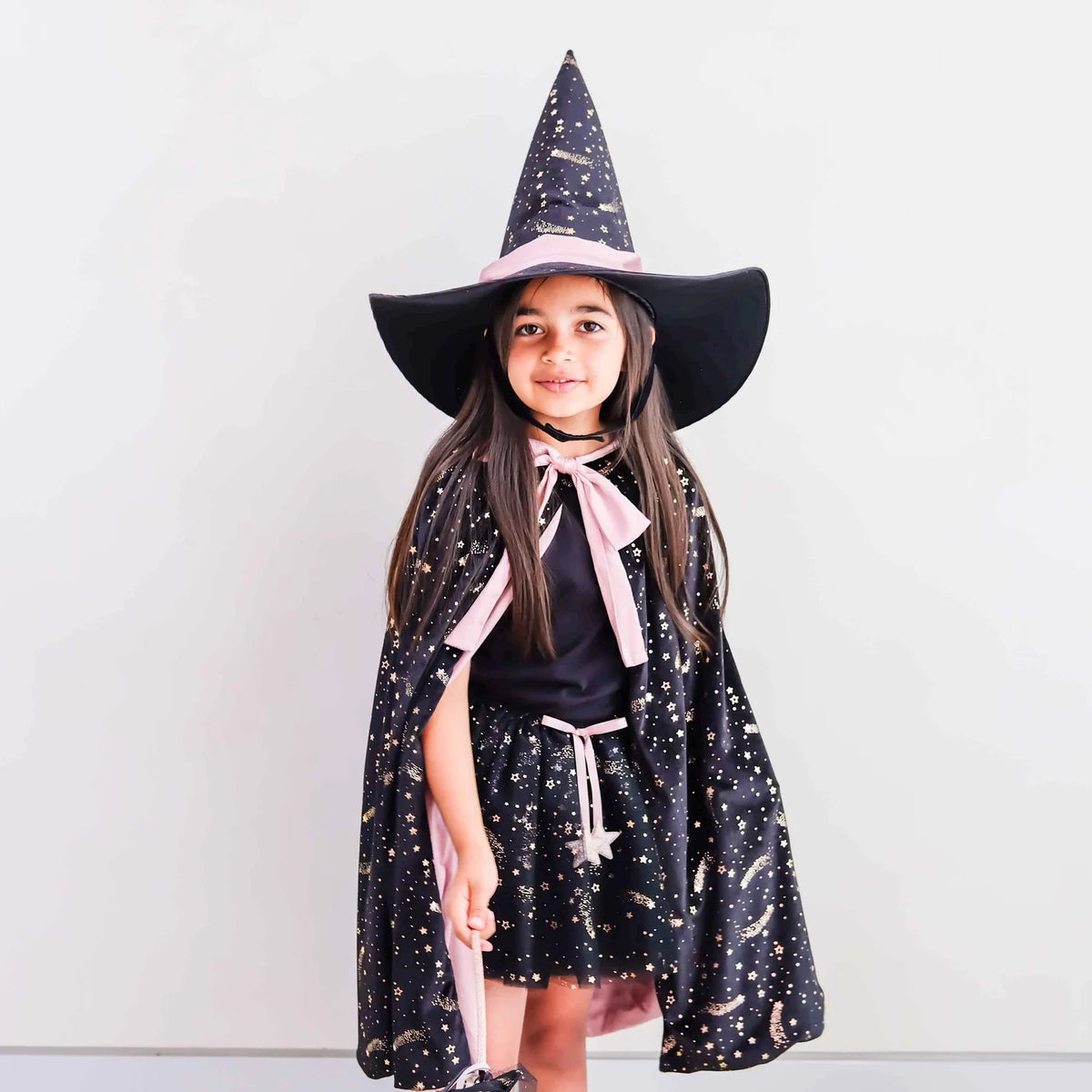 A young girl wearing a black witch costume with a starry pattern, a pink ribbon neck bow, and a matching pointed witch hat, standing against a plain light gray background.