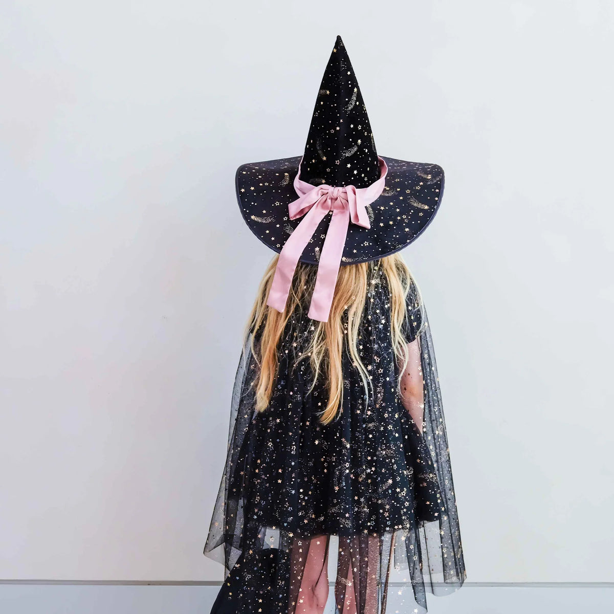 A child viewed from behind wearing a black witch costume with gold speckles, a tall pointed hat, and a pink ribbon bow, standing against a plain light gray wall.