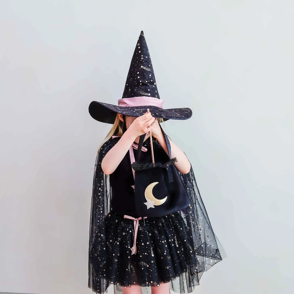 A child wearing a black witch costume with a tall pointed hat, sheer black cape, and tutu skirt, holding a black bag with a gold crescent moon and star. The child is photographed against a plain light gray background.