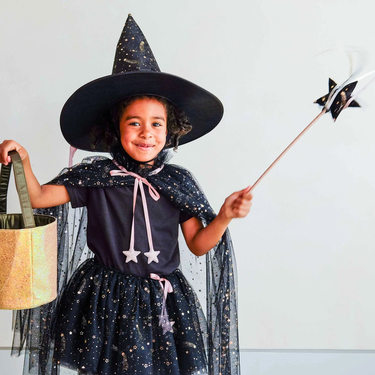 A young child wearing a sparkly black witch costume with a matching pointed hat, sheer cape, and star details, holding a gold glittery bucket and a star wand against a plain light background.