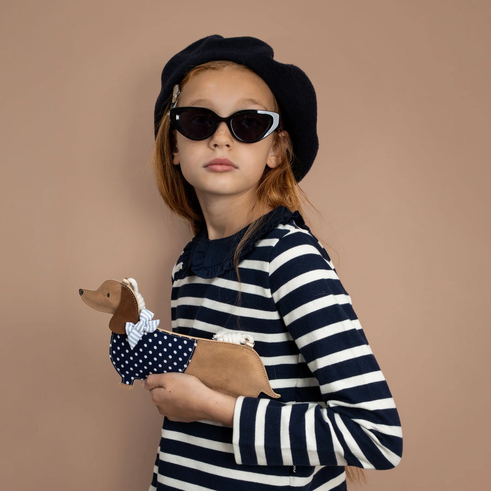 Young girl in a striped outfit holding a plush dog bag against a beige background