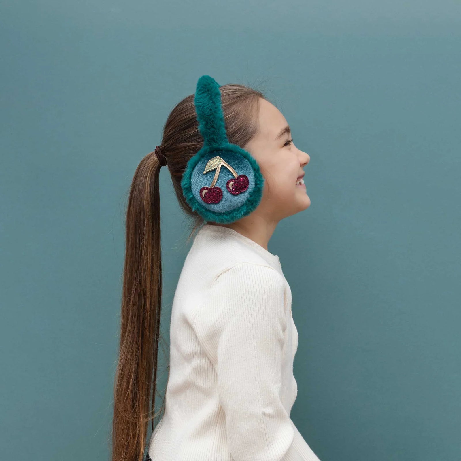 A young girl with a brown ponytail is shown in profile wearing teal fuzzy earmuffs decorated with embroidered cherries, set against a solid teal background.