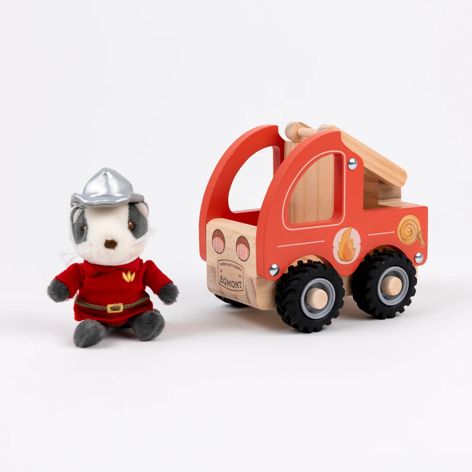 A plush dog firefighter toy sitting next to a wooden fire truck on a clean white background. The dog is wearing a red firefighter suit and silver helmet, while the toy truck is made of natural wood with red-painted panels and black wheels.