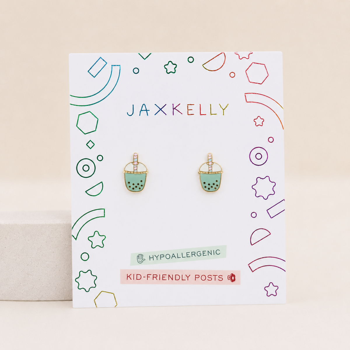 A pair of Jaxx Kelly stud earrings shaped like boba tea cups, displayed on a white branded backing card. The earrings have mint green bodies with black boba dots and gold-toned outlines and straws, set against a light beige background.