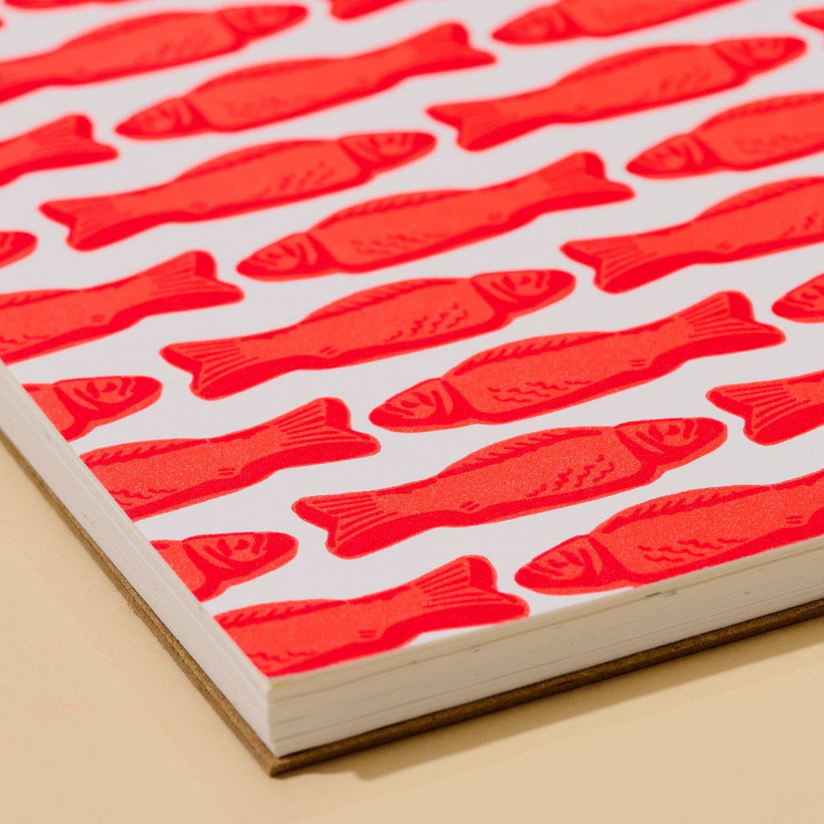 A close-up of a printed sheet or book cover featuring a repeating pattern of bright red fish on a white background, photographed at an angle against a warm beige surface.