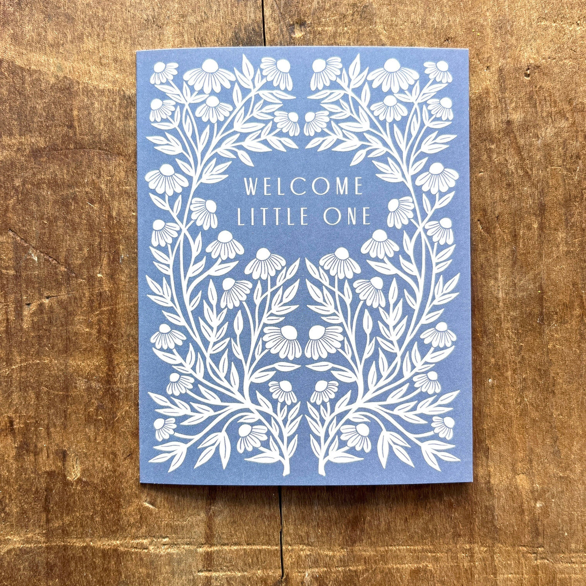 Blue card with white floral design and 'Welcome Little One' text on a wooden surface