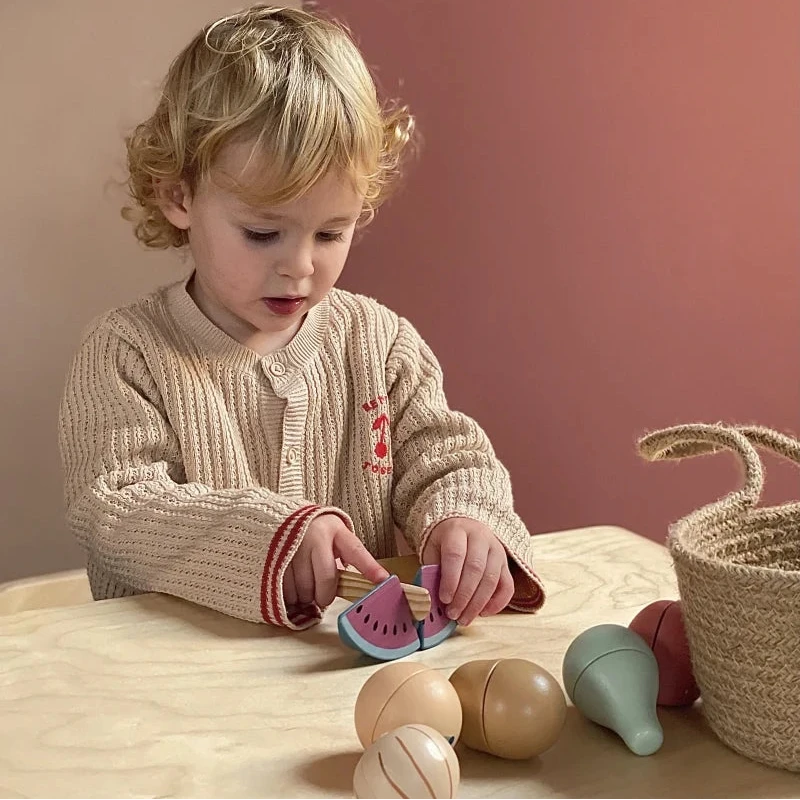 A toddler with curly blonde hair sits at a light wood table, playing with a set of pastel-colored wooden toy fruits and vegetables. The child is wearing a beige knit cardigan with red trim.
