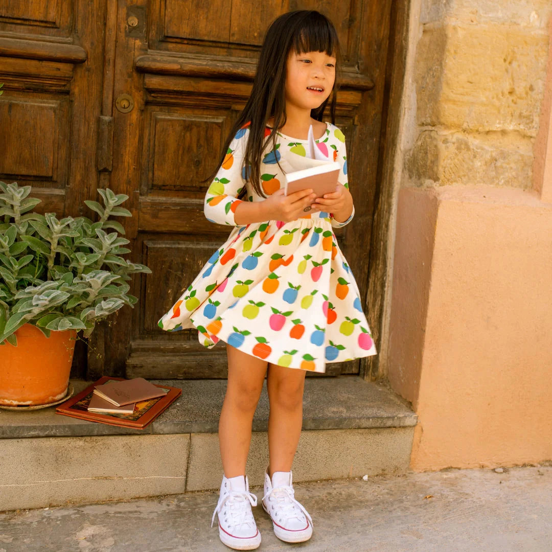 Young girl in a colorful apple print dress standing outside a wooden door.