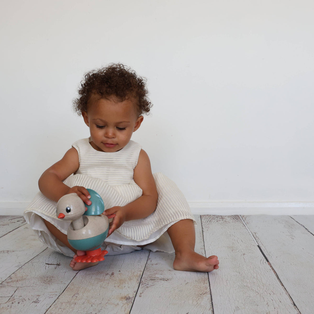 A toddler with curly brown hair sits on a light wood floor against a white wall, holding a chicken toy. The child is wearing a white sleeveless dress.