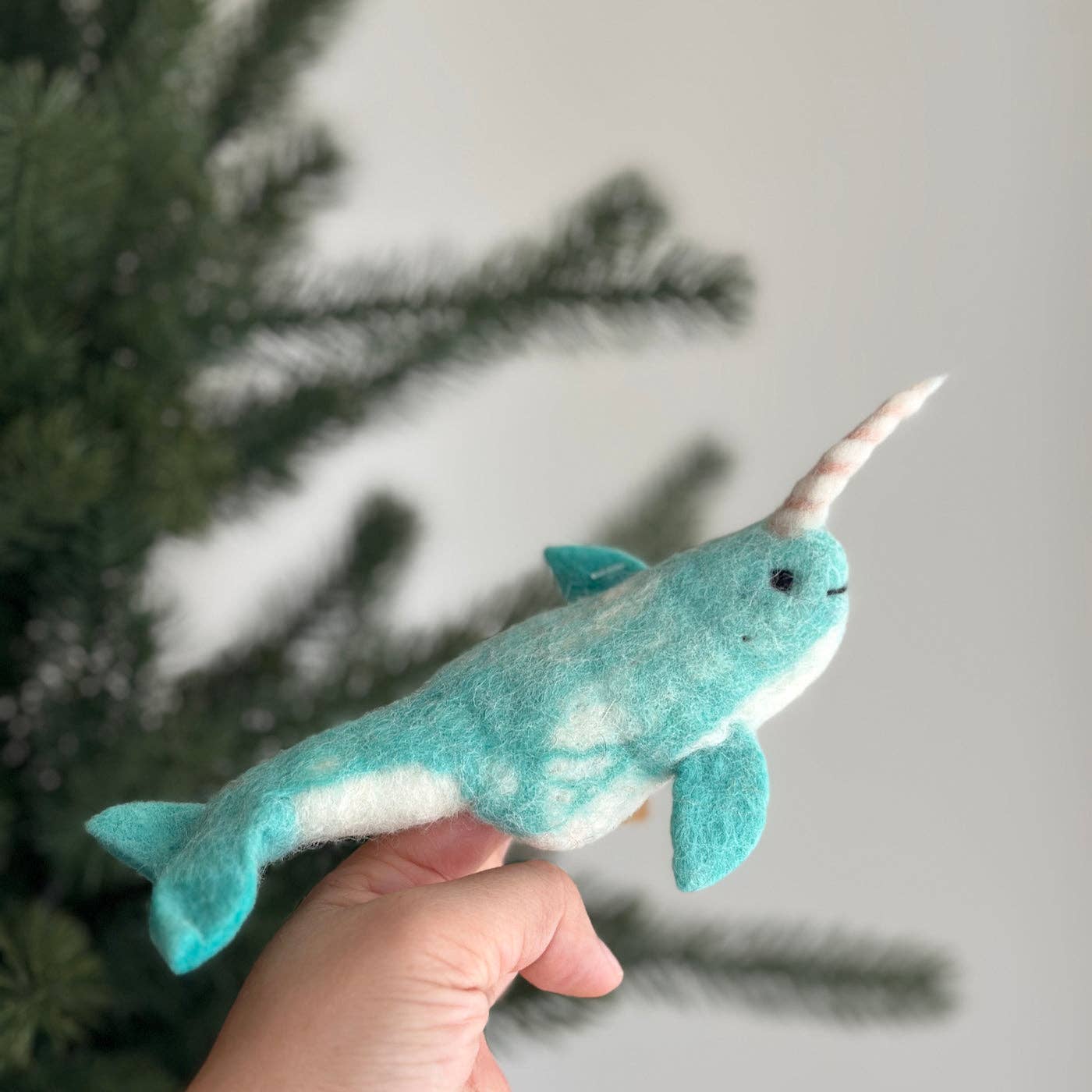Plush toy narwhal held in a hand with a blurred Christmas tree background