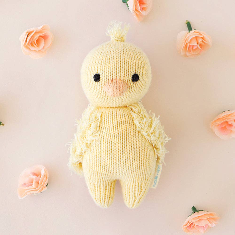 Knitted yellow chick toy surrounded by small pink flowers on a light background