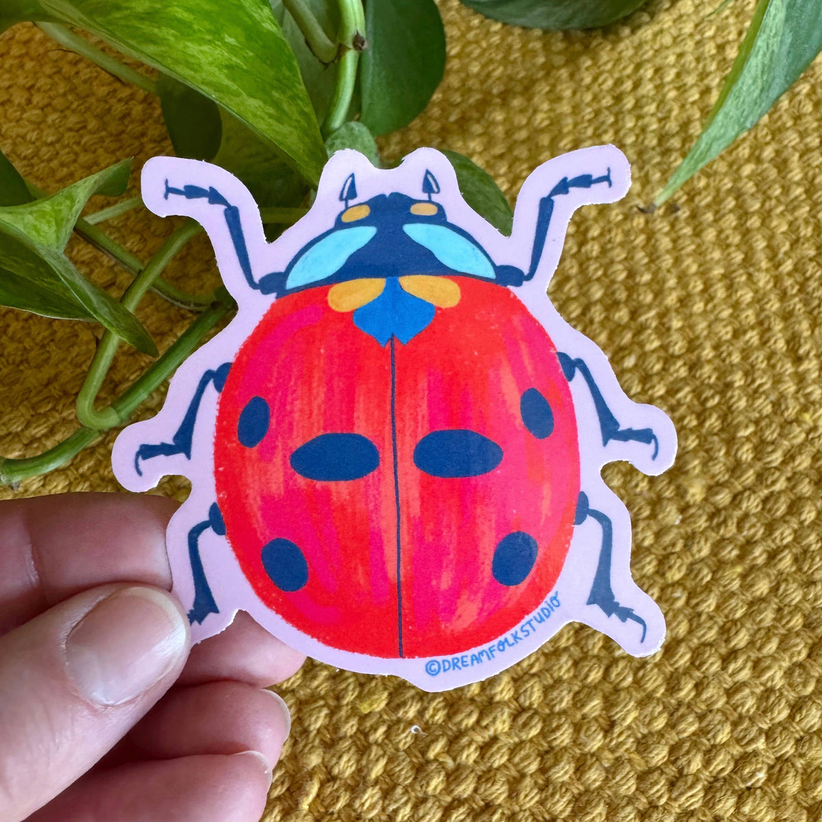 Colorful ladybug sticker held by a hand on a textured yellow surface with green leaves.