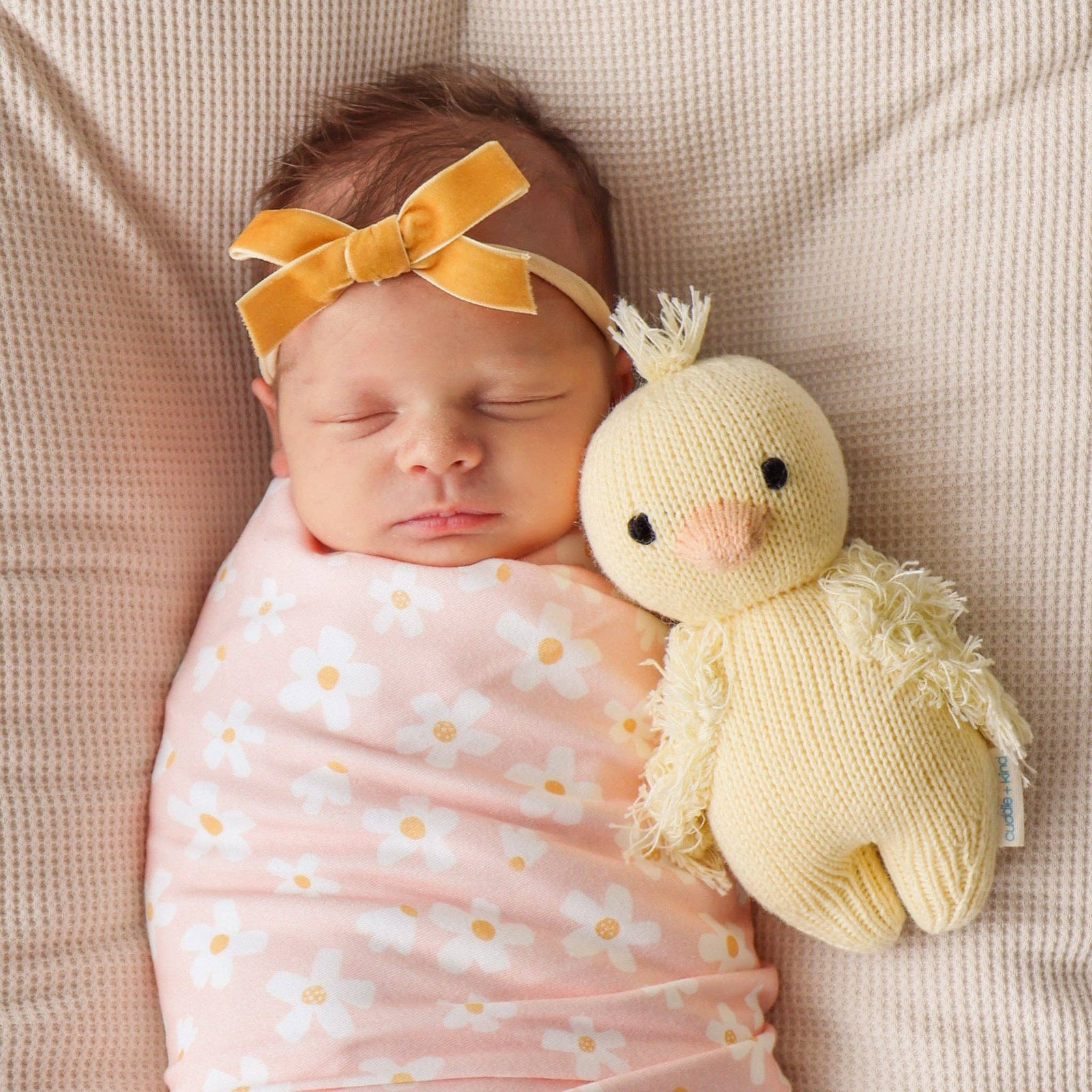 Knitted yellow chick toy surrounded by small pink flowers on a light background