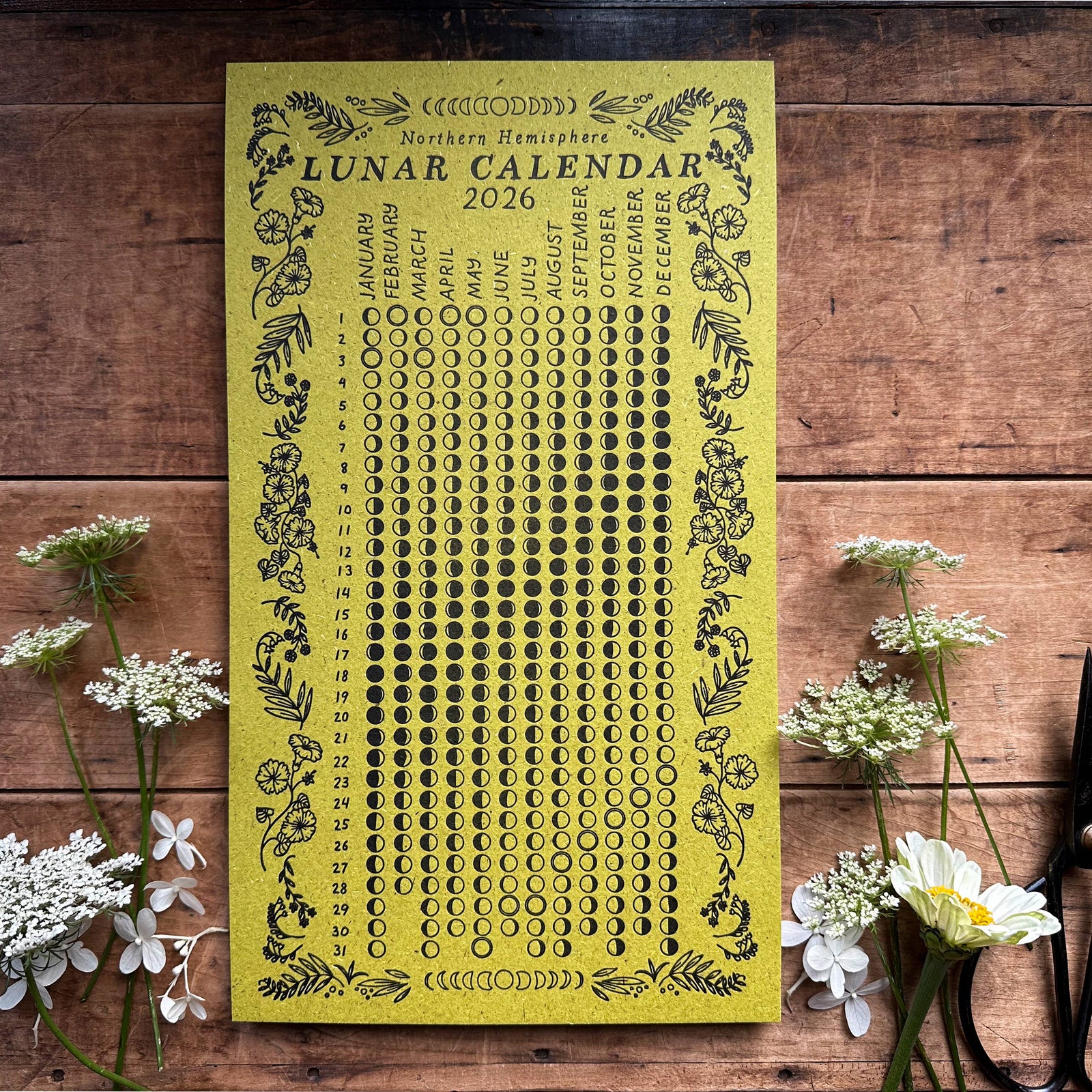 Yellow lunar calendar 2026 on a wooden surface with flowers and scissors.