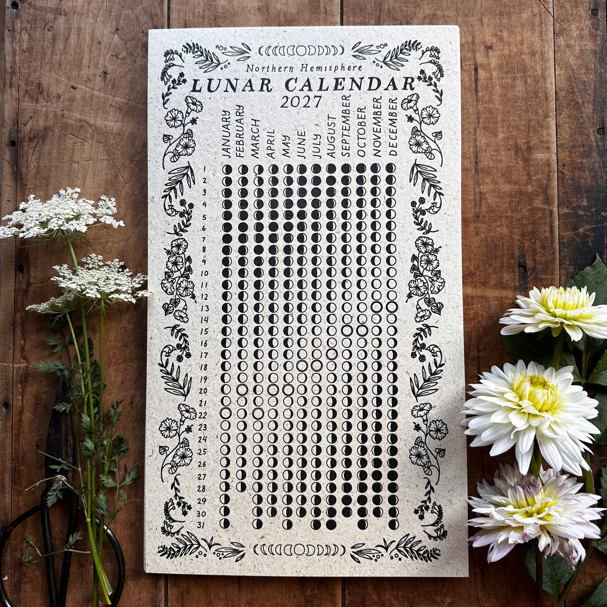 A 2027 Lunar Calendar  printed on a cream-colored cardstock with black ornate illustrations, displayed flat on a rustic wooden background with white flowers and scissors as props.