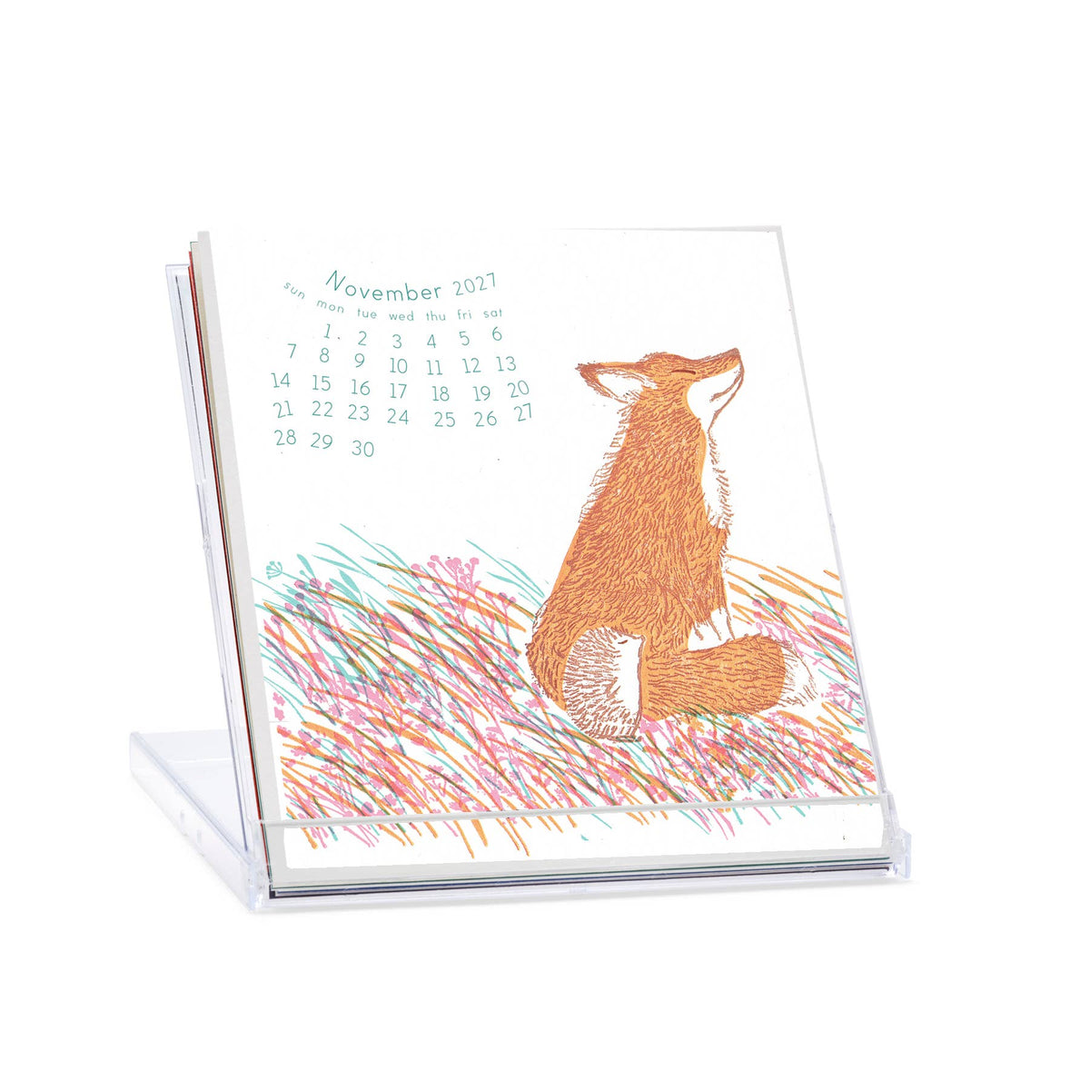 A clear acrylic desk calendar stand holding a printed calendar page with a fox illustration and the date November 2021. The page shows a white background with a brown fox sitting in multicolored grass and a small calendar grid at the top.