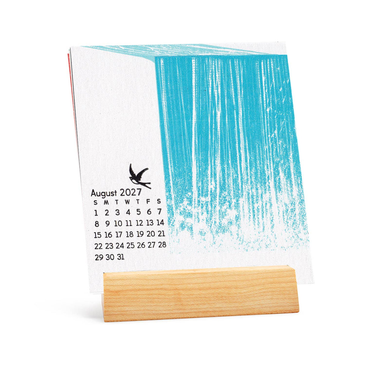 A small desk calendar standing on a light wood block, featuring a white page with a bright turquoise abstract brushstroke design and a printed August 2027 calendar grid with a bird icon.