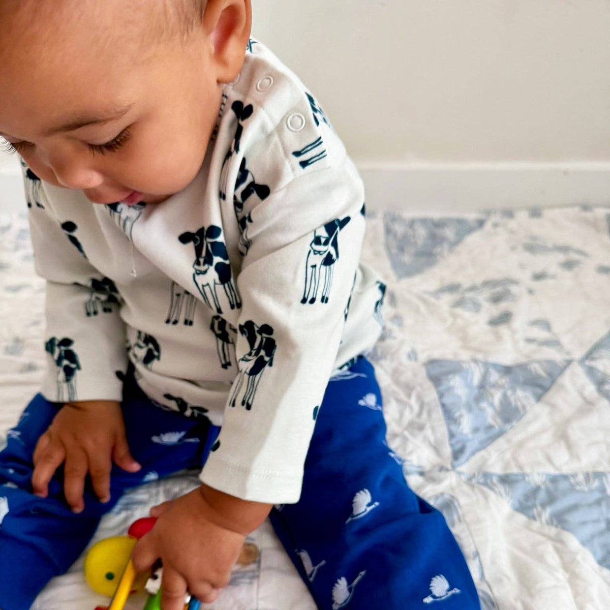 A baby sitting on a quilted bed, wearing a white long-sleeve top with black cow prints and blue pants with white cow prints, holding a small toy.