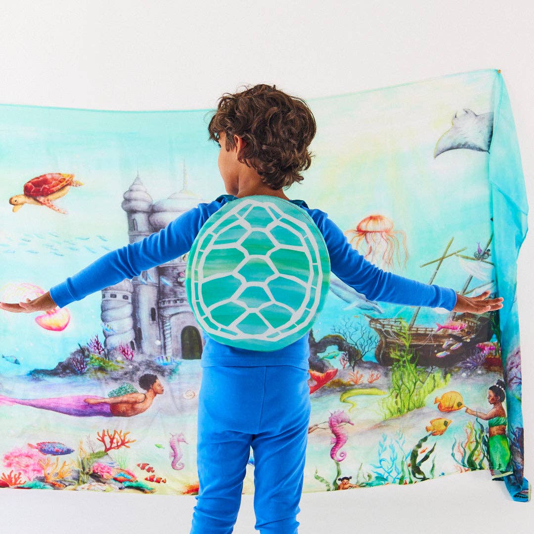 A child stands with arms outstretched, wearing a bright blue outfit and a large turtle-shaped back cover that displays a colorful underwater scene with a castle, sea turtle, jellyfish, and coral. A matching printed fabric backdrop hangs behind them, showing the full illustrated scene.