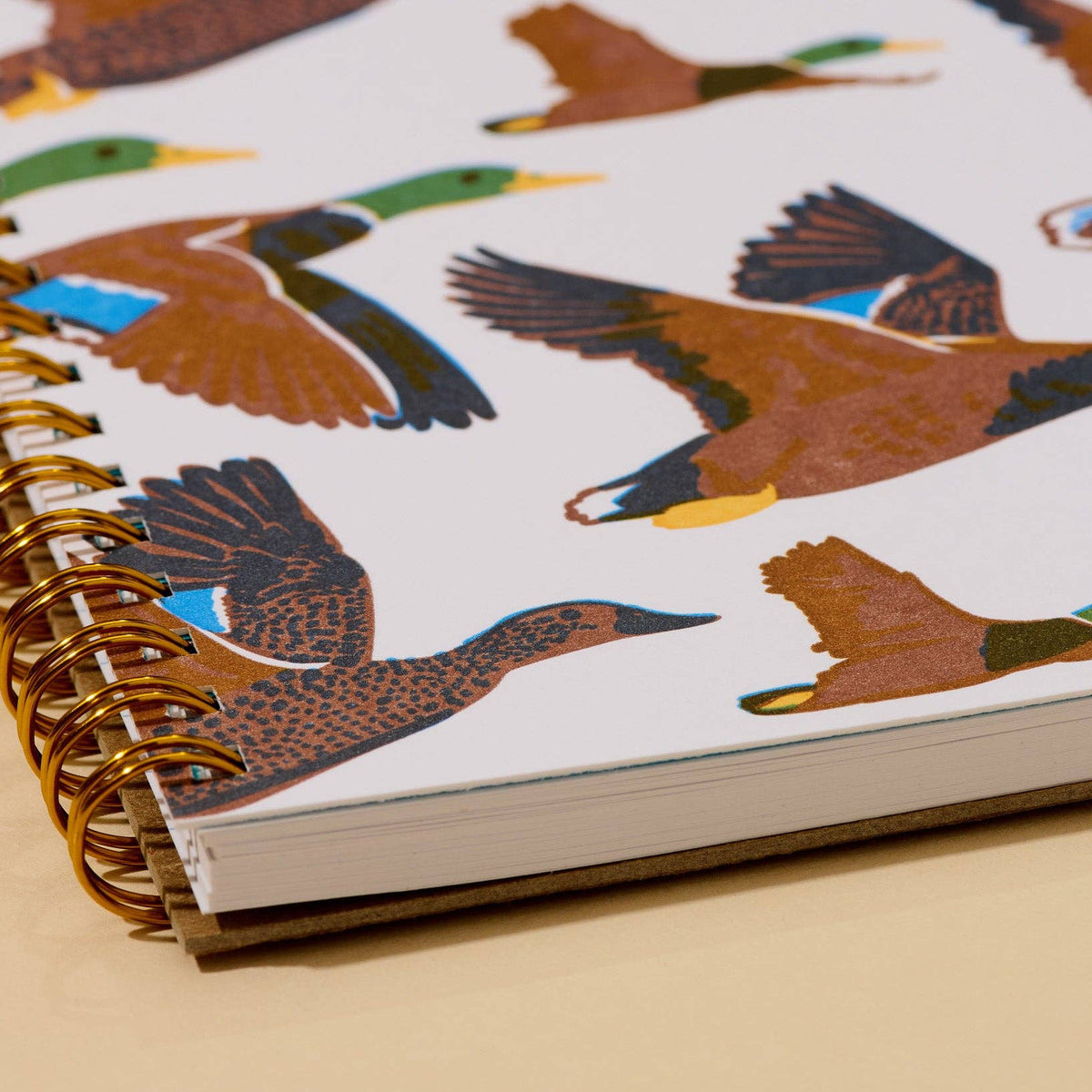 A close-up of a spiral-bound notebook with a white cover printed with colorful duck illustrations, bound with a gold-tone spiral, on a beige surface.