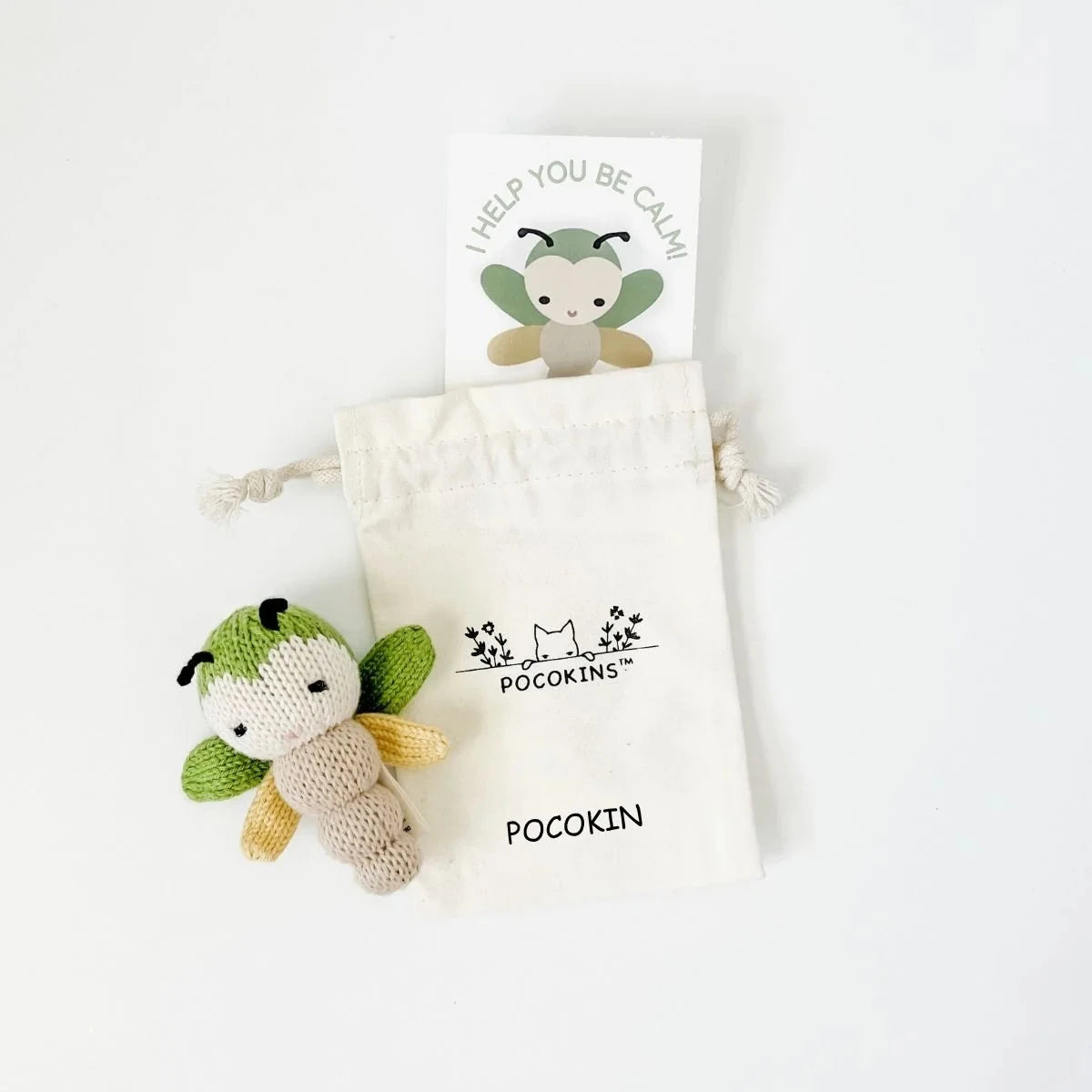 A small knitted dragonfly toy with green wings and a beige body, shown next to a cream drawstring pouch printed with the brand name 'Pocokins' and a matching card on a white background.