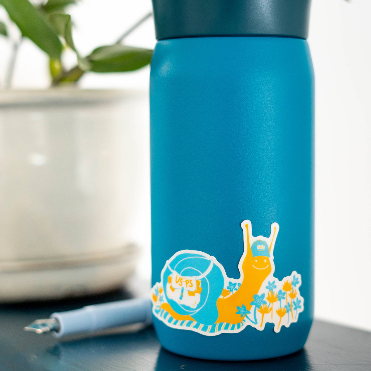 A blue insulated tumbler with a dark blue lid, featuring a cartoon snail graphic in yellow, orange, and white near the bottom, shown on a tabletop with a blurred potted plant in the background.