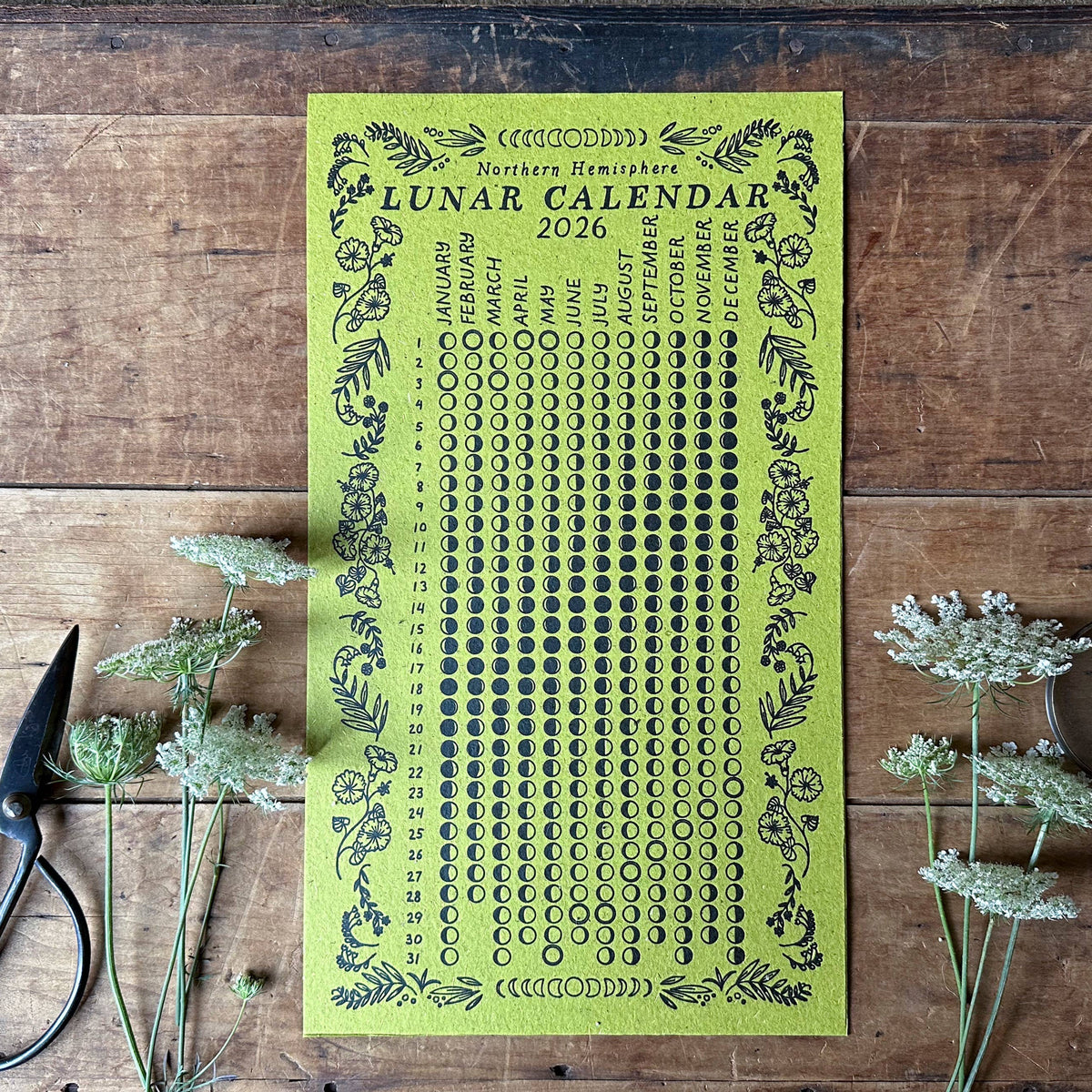 Green lunar calendar 2026 on a wooden surface with scissors and flowers.