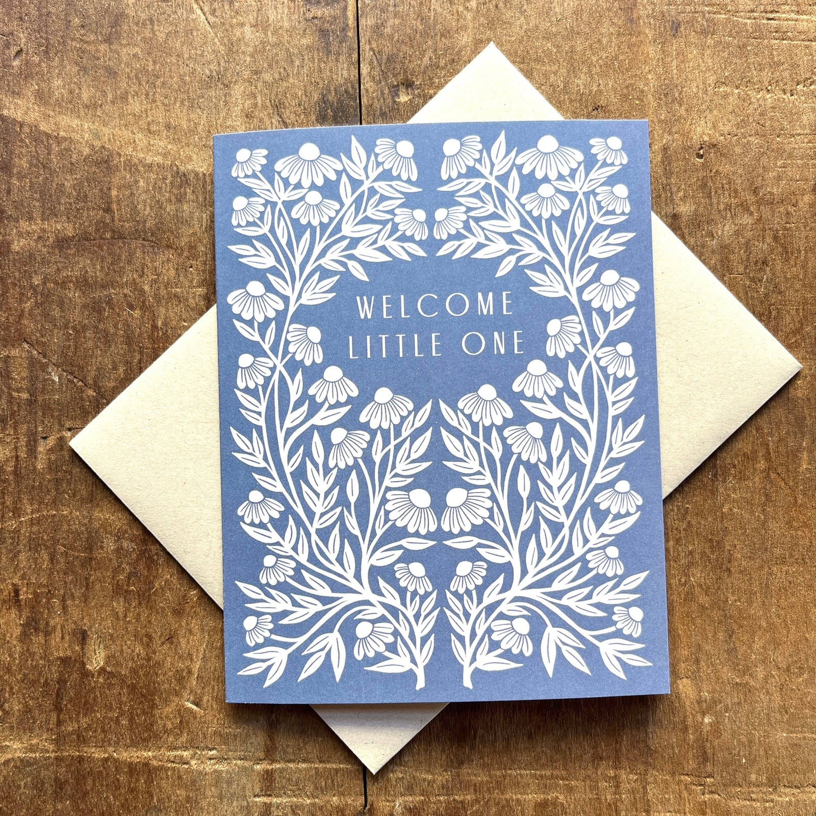 Blue card with white floral design and 'Welcome Little One' text on a wooden surface