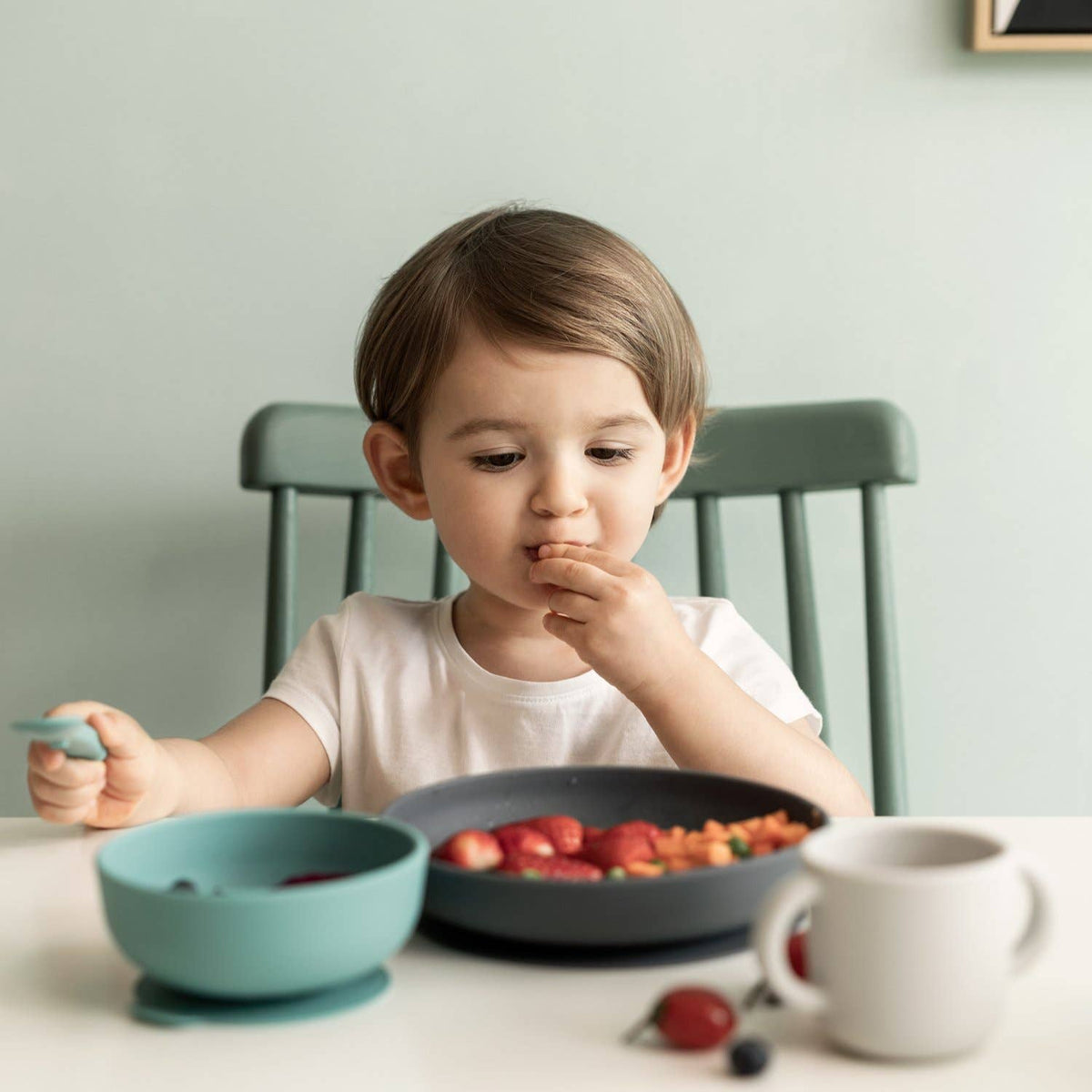 Child eating at a table with a bowl of fruit and a cup.