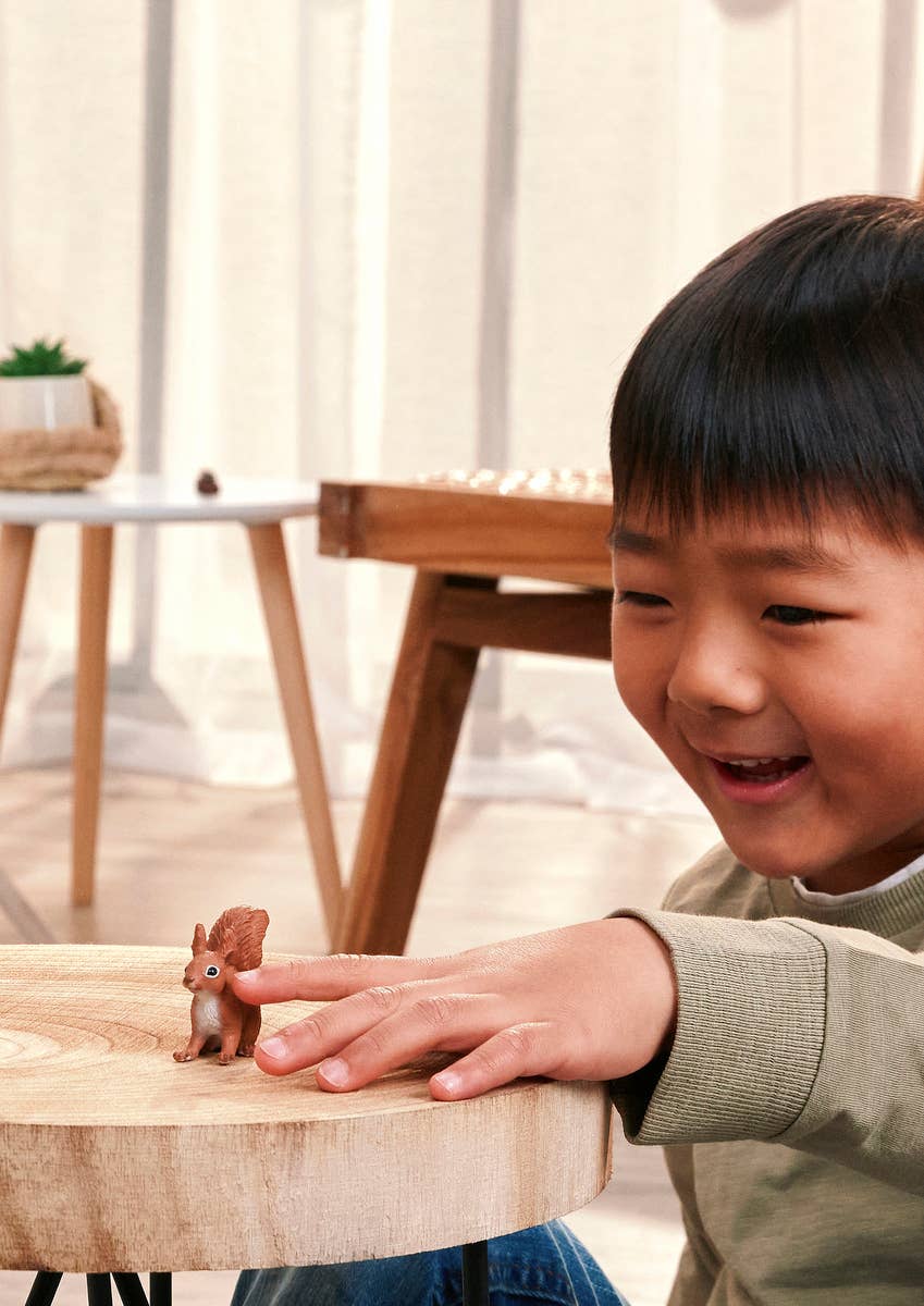 A young boy smiling and touching a small brown squirrel figurine sitting on a round wooden table in a bright indoor setting.