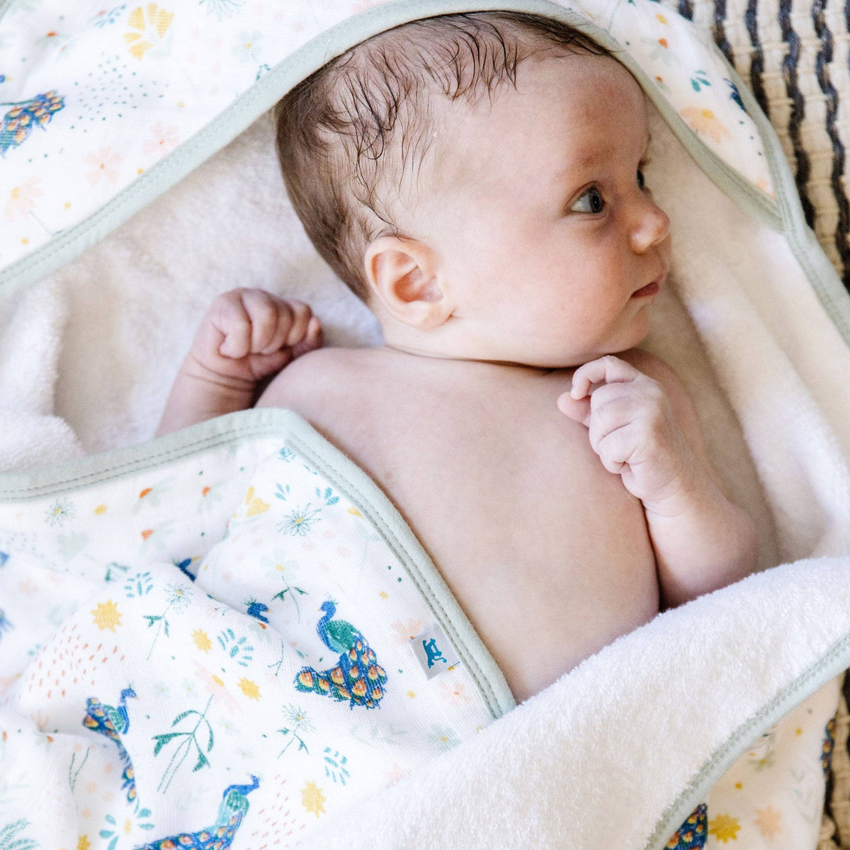 A close-up of a baby wrapped in a soft, cream-colored towel with a colorful peacock and floral print and a plush cream lining.