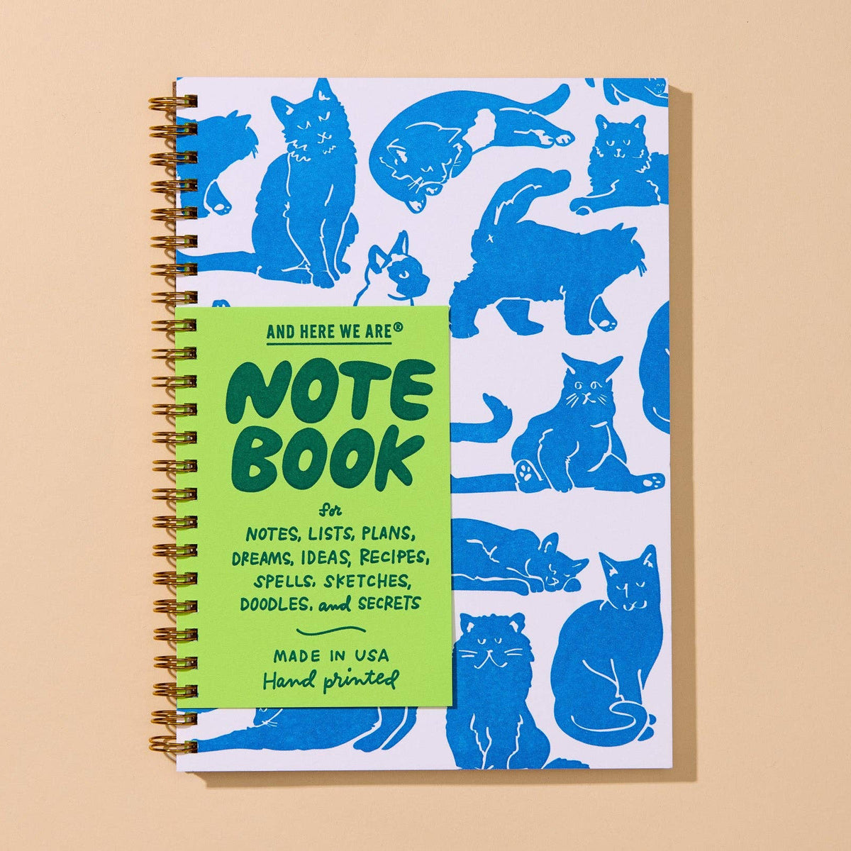 A spiral-bound notebook with a white cover decorated with blue cat illustrations and a bright green front cover that reads 'AND HERE WE ARE! NOTEBOOK'.