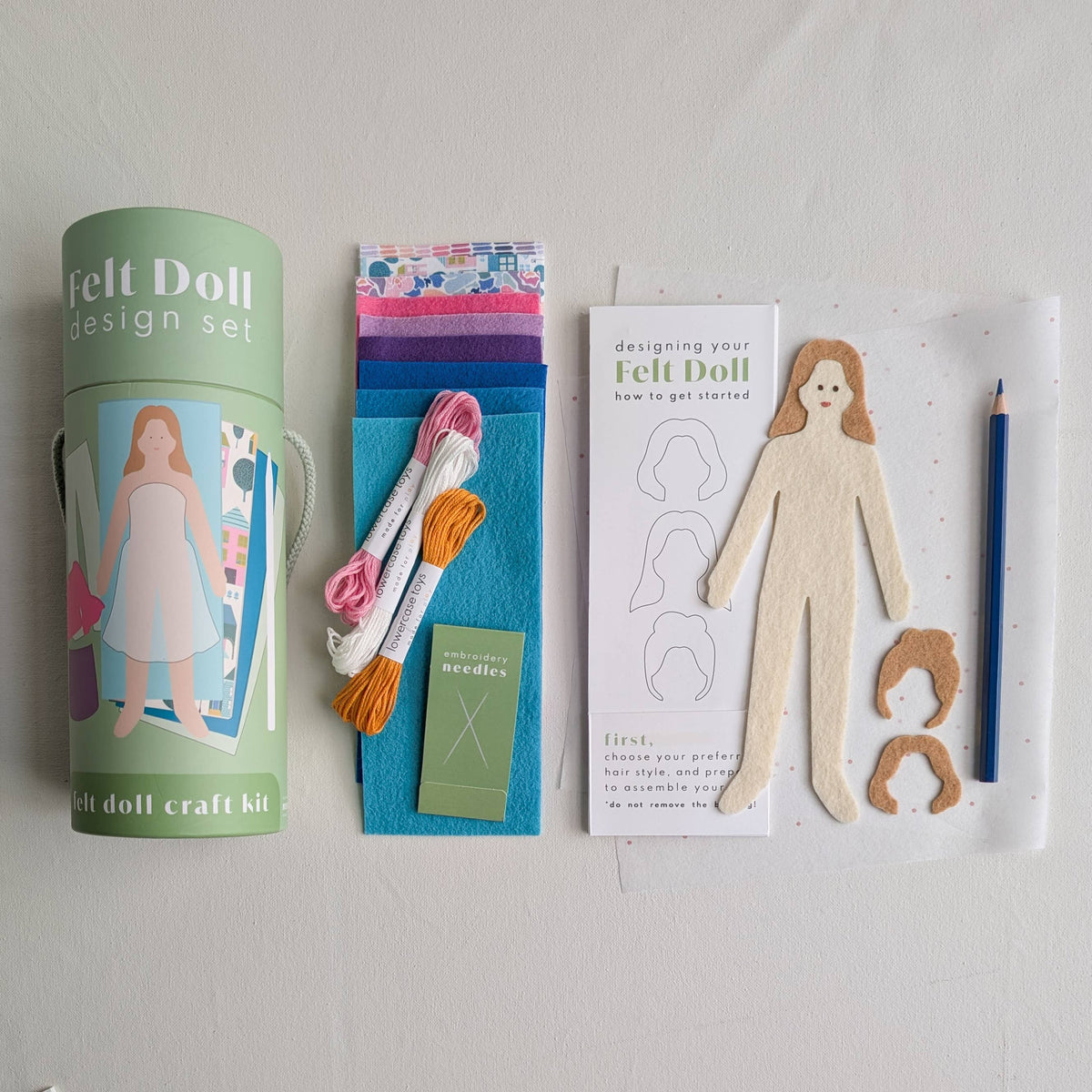 Felt doll design set with packaging, felt pieces, needles, and a paper doll template on a white background.