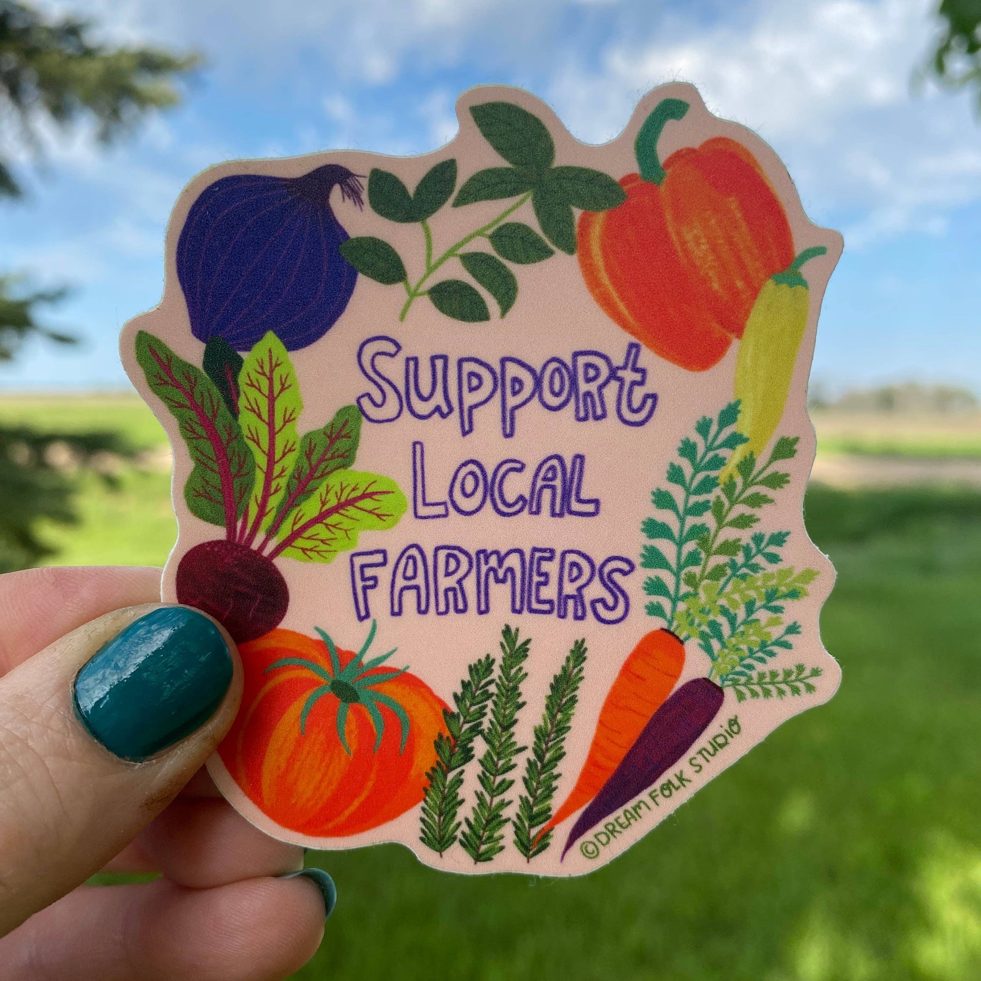 Sticker with 'Support Local Farmers' text and vegetables held by a hand outdoors.
