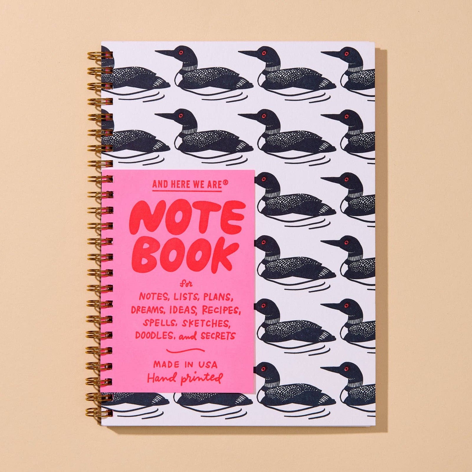 A spiral-bound notebook with a white cover printed with a repeating black-and-white duck pattern, featuring a bright pink front cover with red text reading 'NOTE BOOK' and smaller text below. The notebook is photographed on a warm beige background.