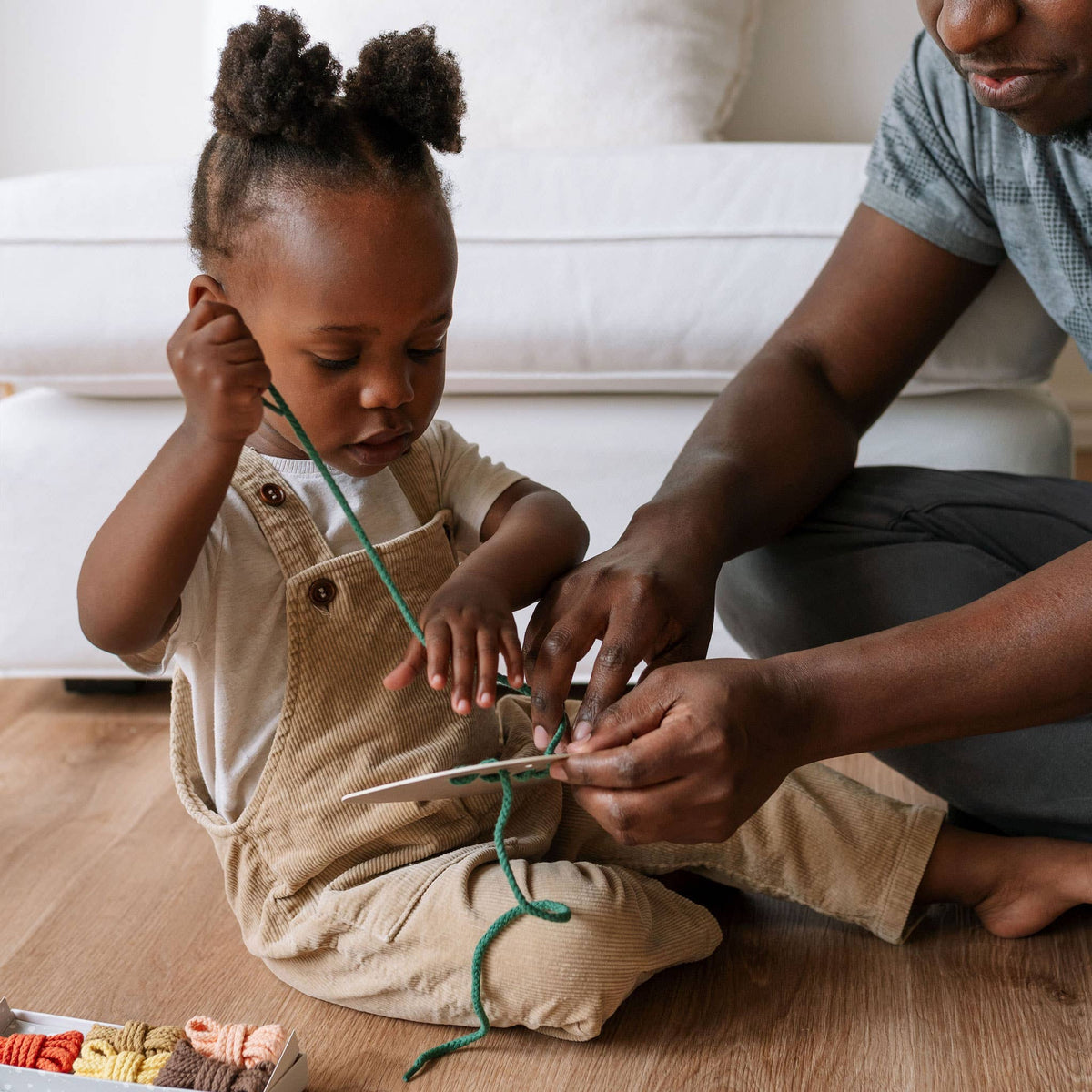 Child and adult sitting on a wooden floor, engaging in craft activity with yarn.