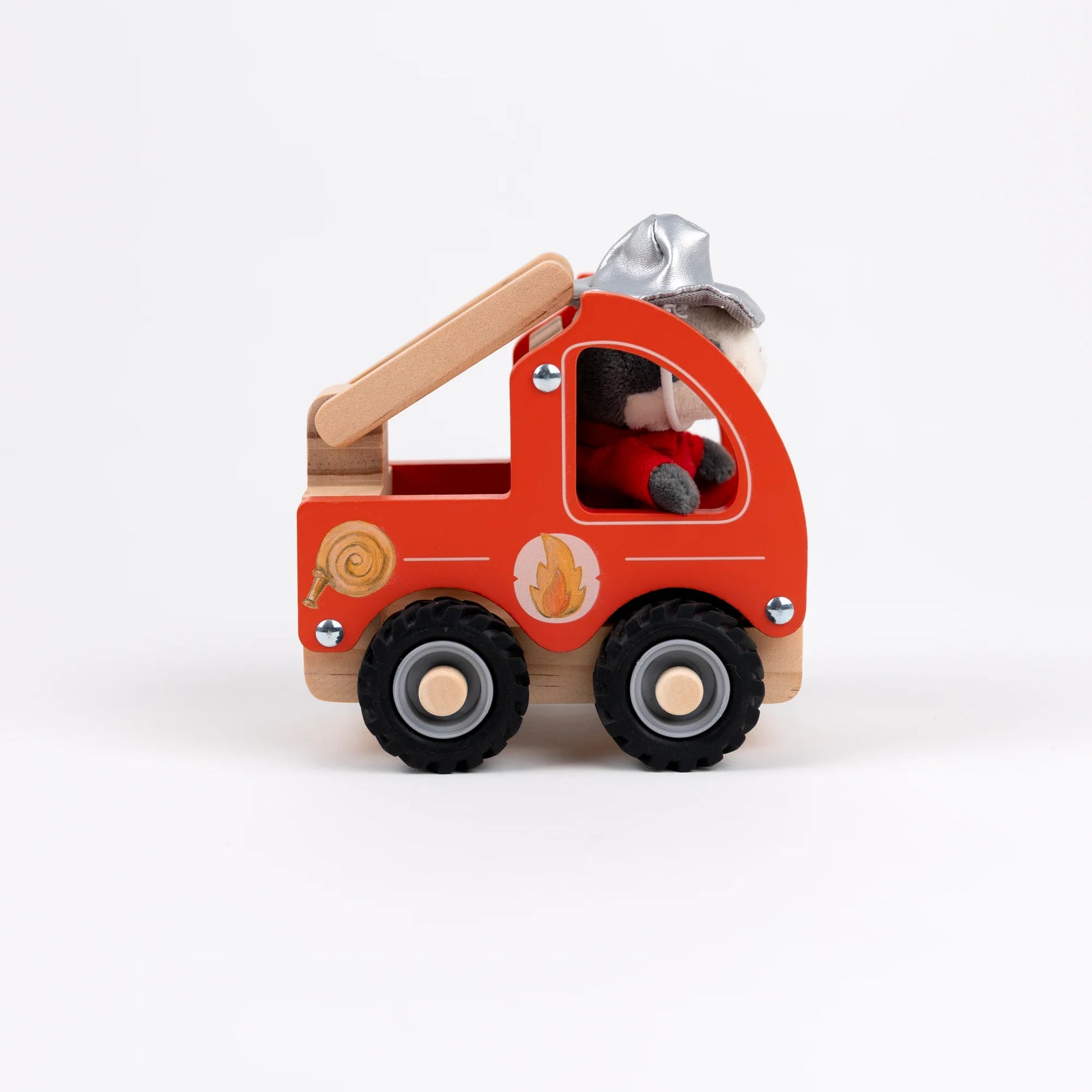 A plush dog firefighter toy sitting next to a wooden fire truck on a clean white background. The dog is wearing a red firefighter suit and silver helmet, while the toy truck is made of natural wood with red-painted panels and black wheels.