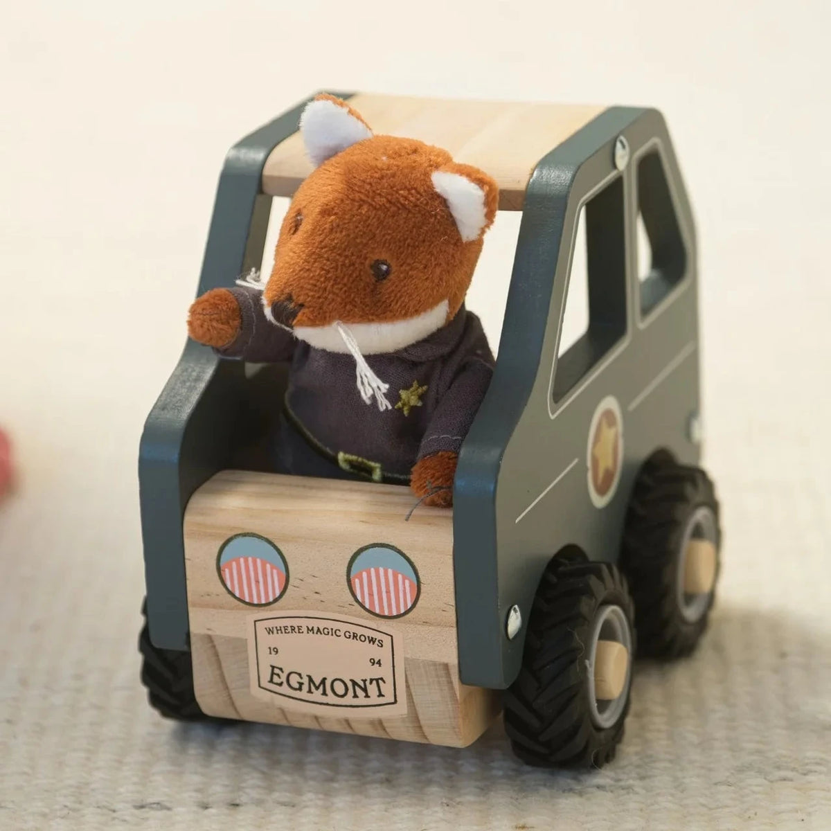 A small plush toy fox sitting inside a wooden toy car, photographed on a light beige textured surface.