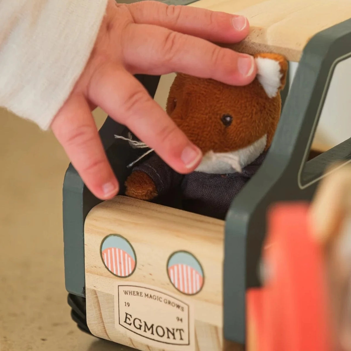 A close-up of a child's hand placing a small brown plush toy inside a wooden toy car. The toy car has a light natural wood body, dark gray side panels, and a front badge that reads 'EGMONT'.