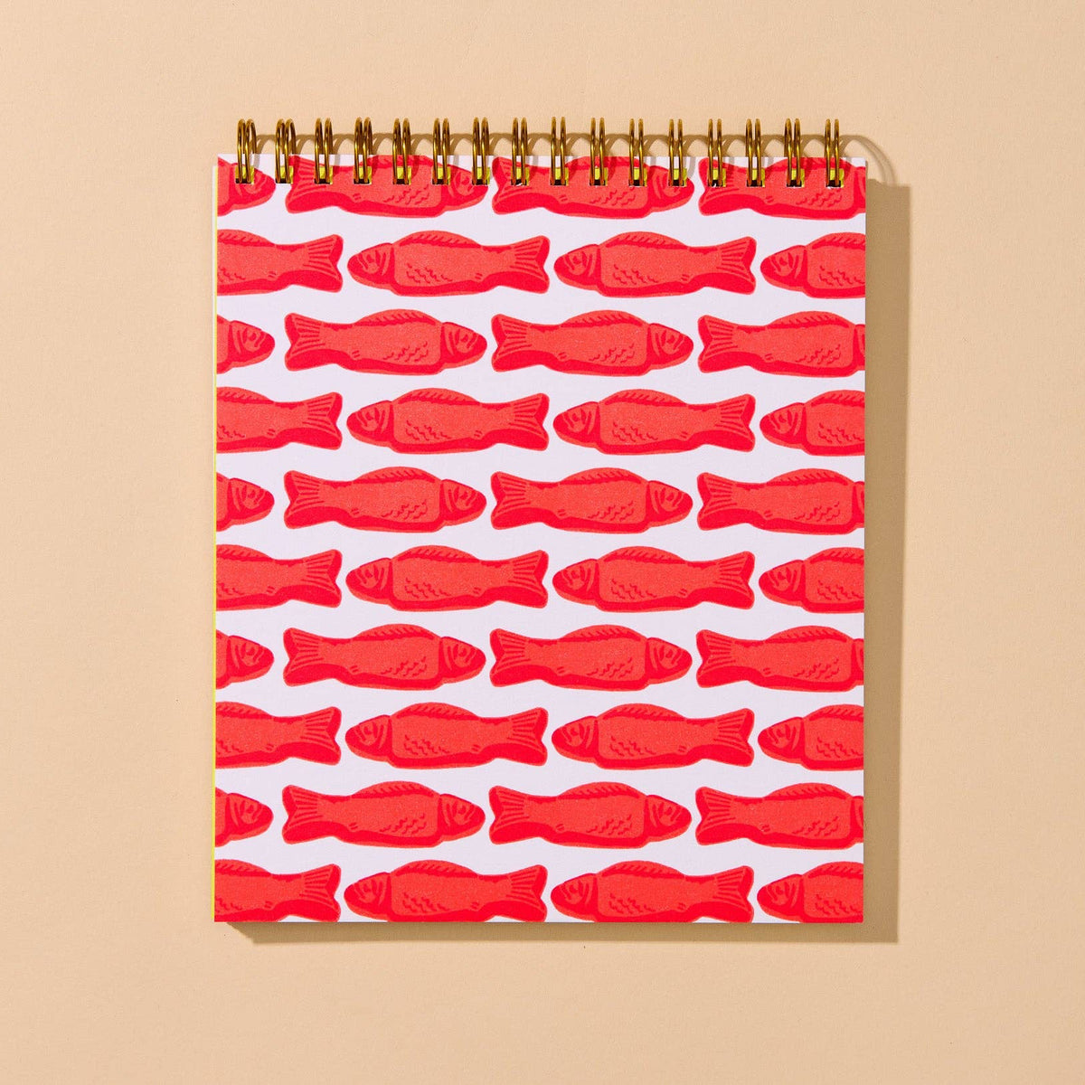 A spiral-bound notebook with a white cover decorated with a repeating pattern of red fish, displayed on a warm beige background.