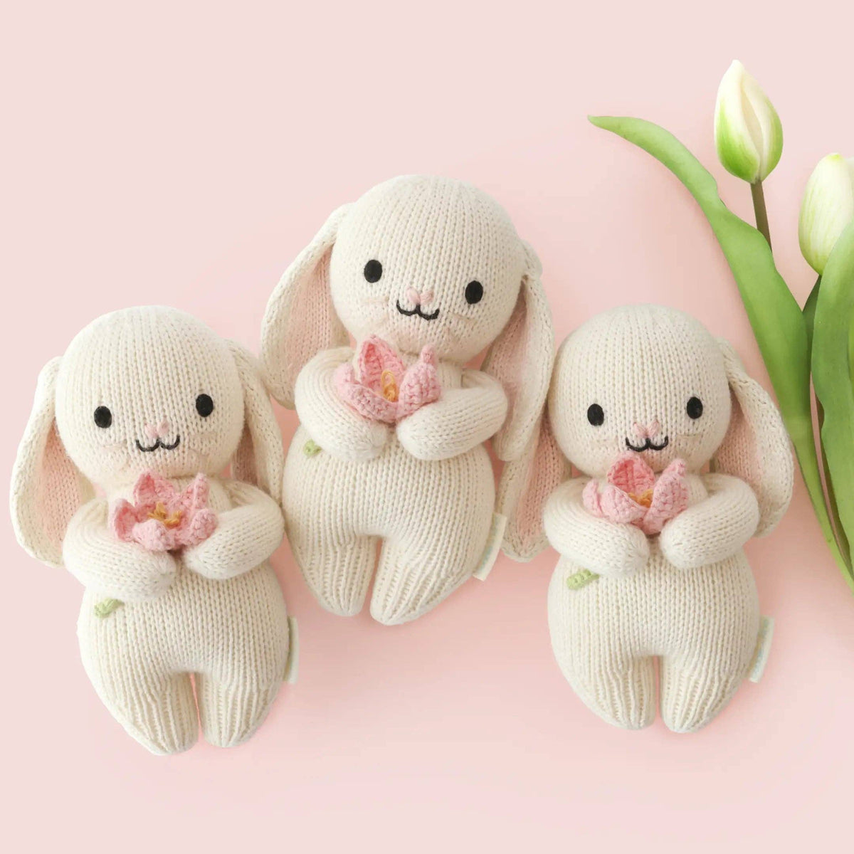 Three cream-colored knitted bunny dolls with black embroidered eyes and mouths, each holding a small pink flower, arranged on a soft pink background with tulip stems at the side.