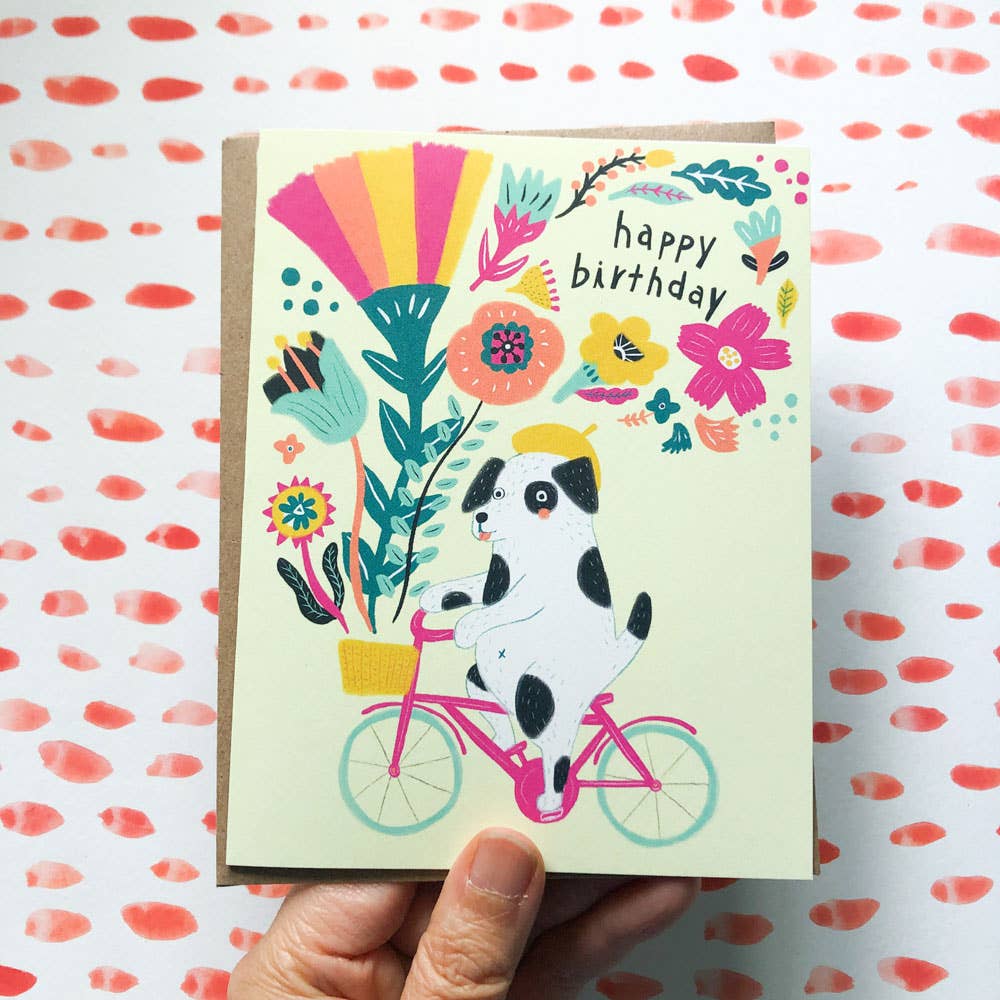 Birthday card with a dog on a bike illustration and colorful flowers on a patterned background