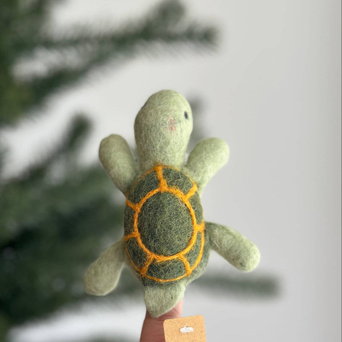 A small felted wool turtle finger puppet held up by a hand, with a light green body, dark green shell, and orange stitched lines, against a softly blurred background of pine branches and a light wall.