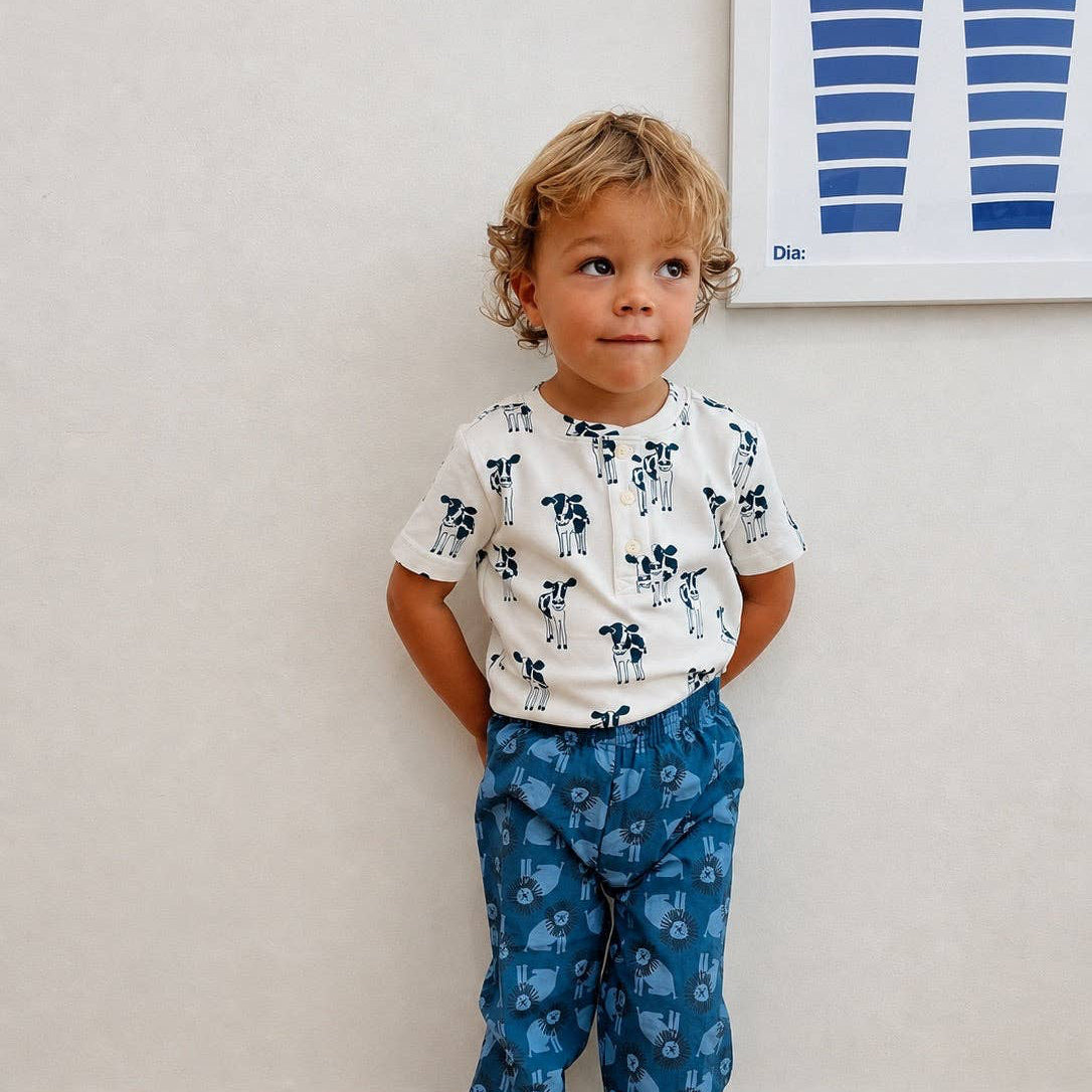 A toddler stands barefoot against a light beige wall wearing a short-sleeve white t-shirt with a blue cow print and blue patterned pants.