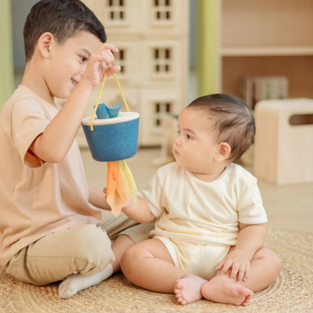 Two children playing with a blue bucket and colorful toy indoors.