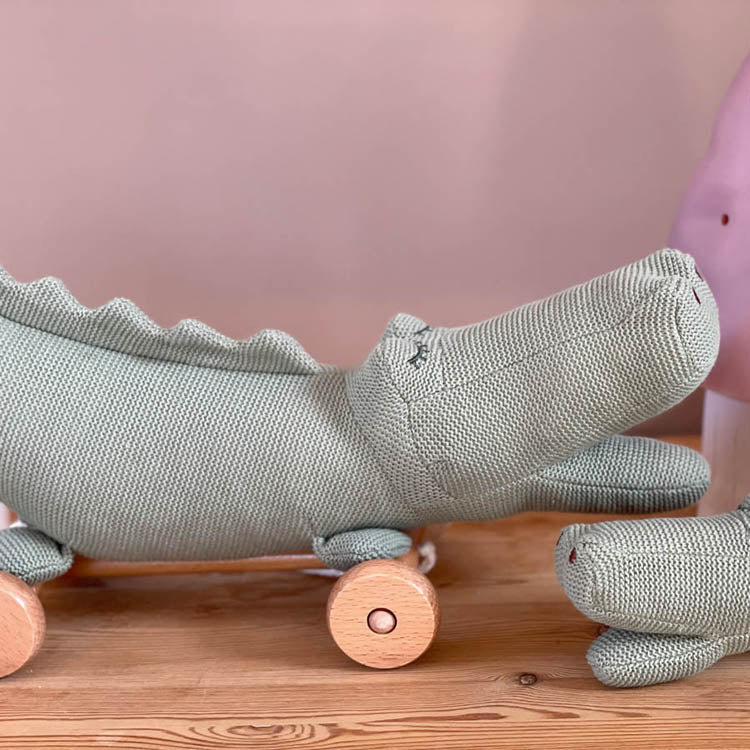 Green toy alligator on wheels next to a pink mushroom on a wooden surface with a pink background
