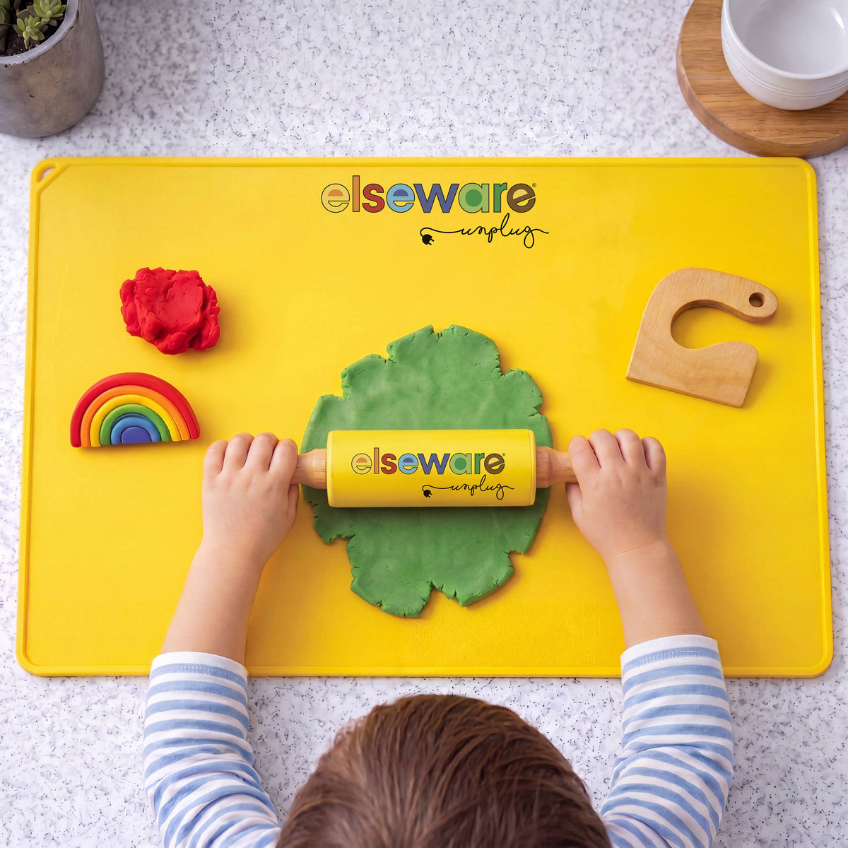 A child's hands and head are visible as they use a yellow Elseware Unplugged rolling pin to flatten green playdough on a bright yellow mat. The scene includes colorful playdough pieces, a wooden letter C, and a speckled countertop background.