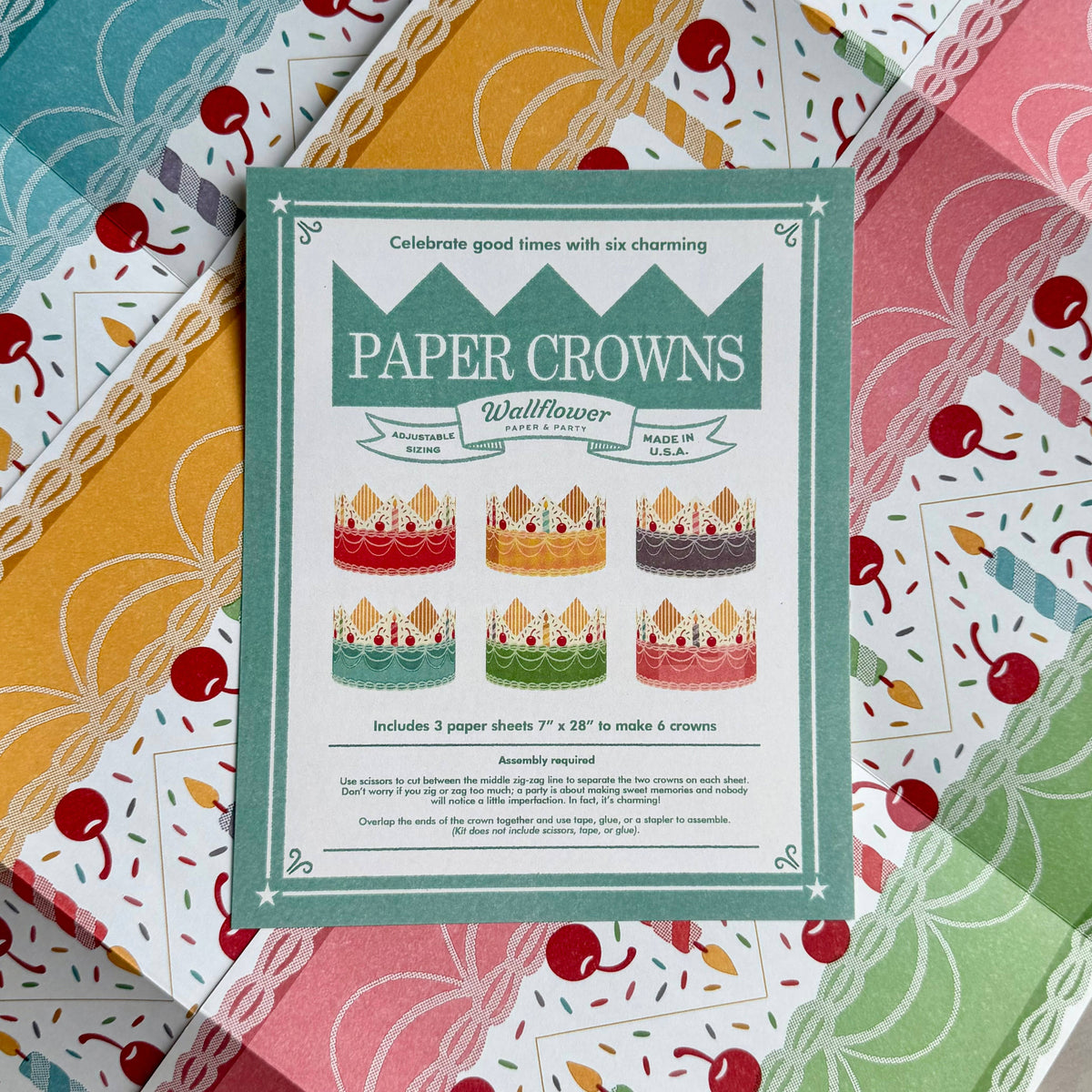 A flat-lay of a 'Paper Crowns' wallflower card set by Wallflower, featuring a central mint green instruction card surrounded by six colorful patterned paper sheets with cherries and stripes.