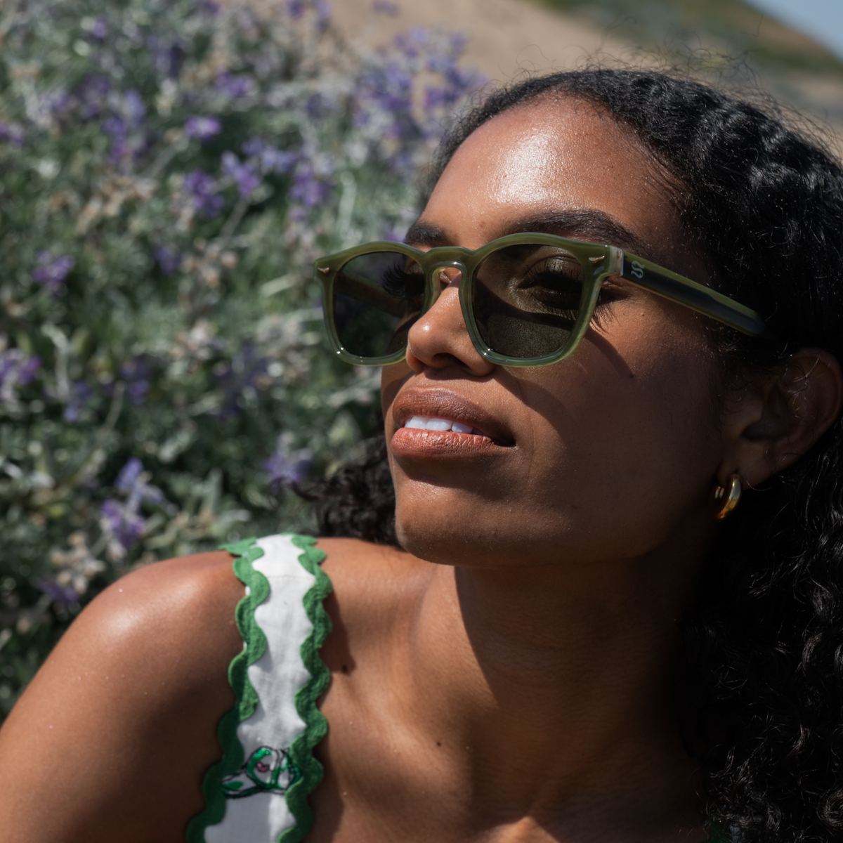 A close-up lifestyle shot of a woman outdoors wearing olive green sunglasses with dark lenses and a white top with green scalloped trim, set against a background of purple flowers and green foliage.